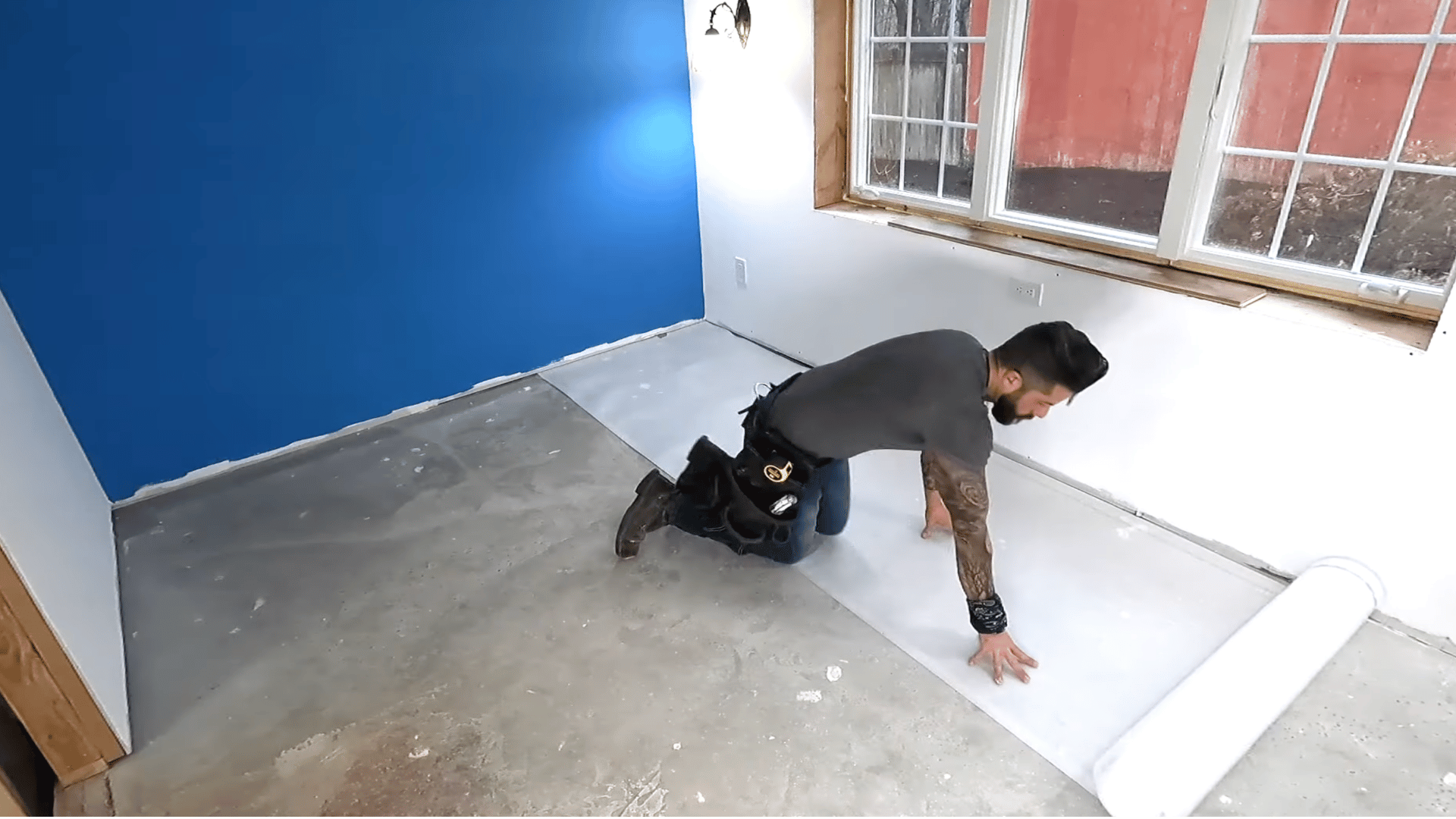 A person kneeling and unrolling underlayment on the floor in preparation for laminate flooring installation, with a blue wall in the background.