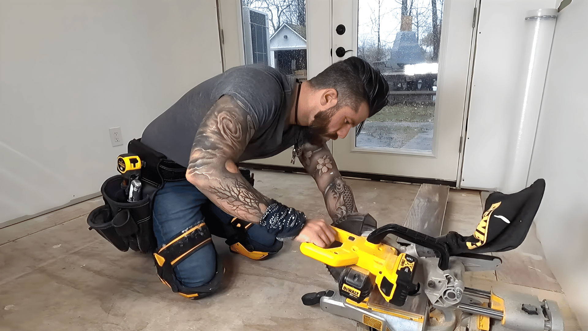 A person using a chop saw to cut laminate planks for installation, with tools on a tool belt and a work area with a floor partially prepared