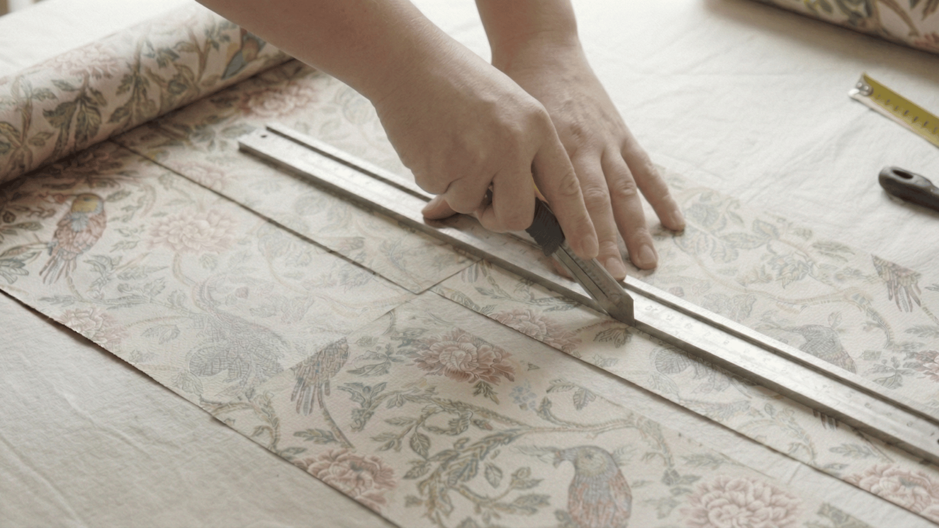 A person using a utility knife and metal ruler to precisely cut floral-patterned wallpaper.