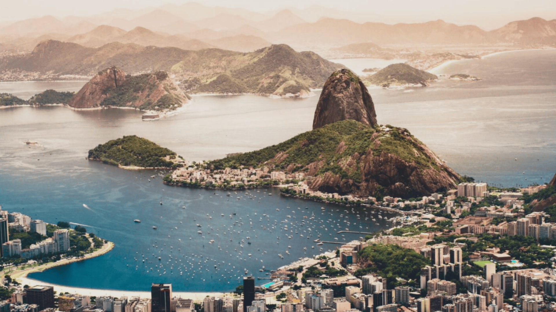 Aerial view of Rio de Janeiro showing Guanabara Bay, Sugarloaf Mountain, green hills, boats on blue water, and dense city buildings.