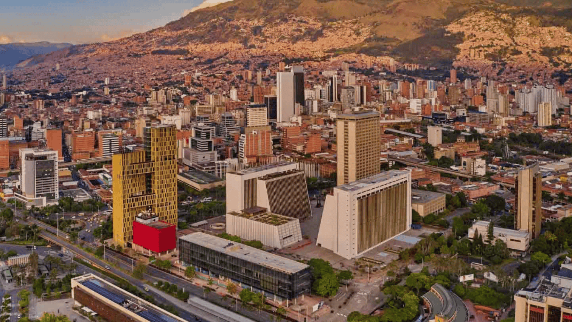 Aerial view of a dense city with modern high rises and wide roads, backed by sprawling hillside neighborhoods under warm light.