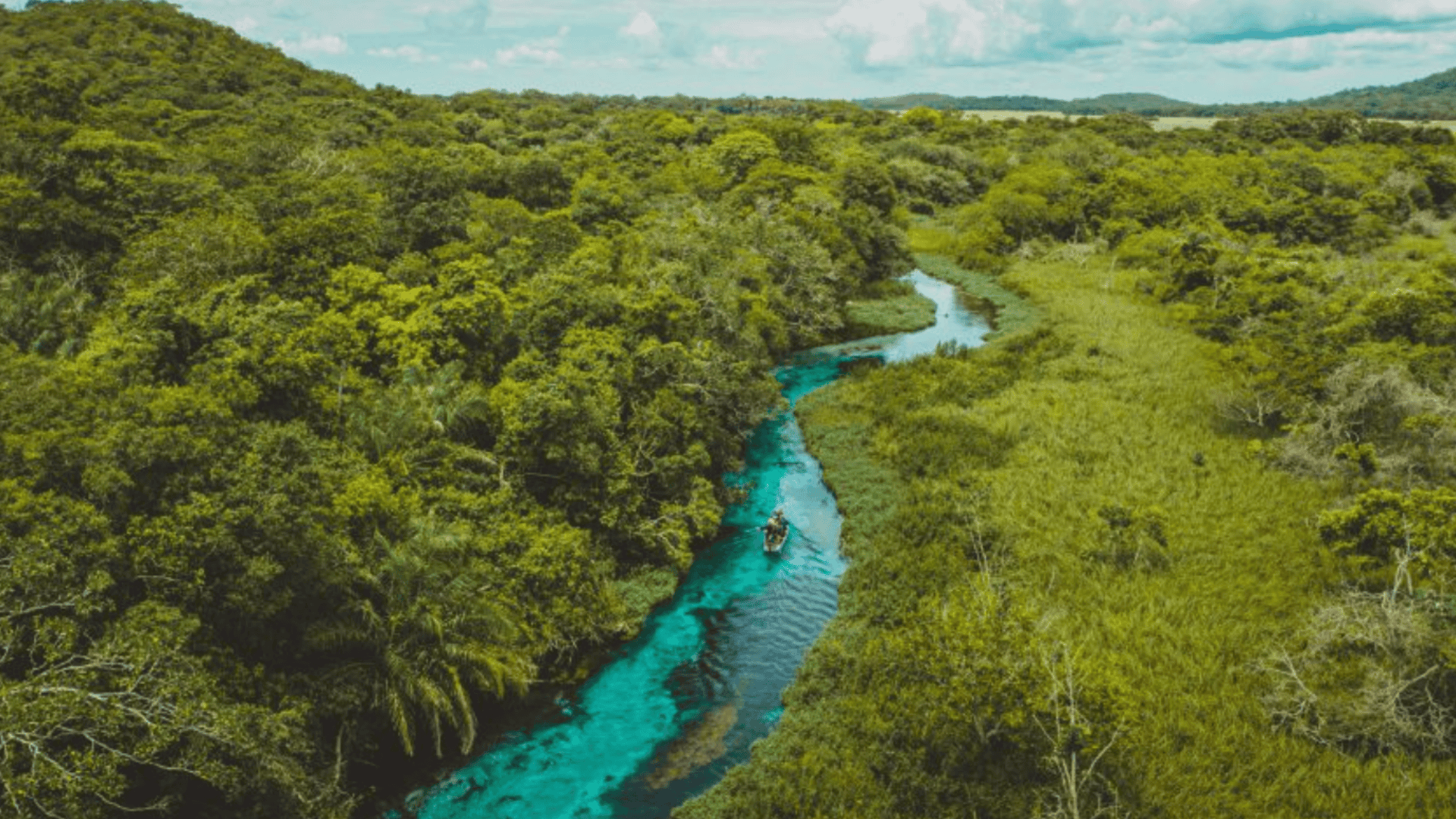 Aerial view of a winding turquoise river through lush green forest and wetlands, with a small boat moving under a cloudy sky.
