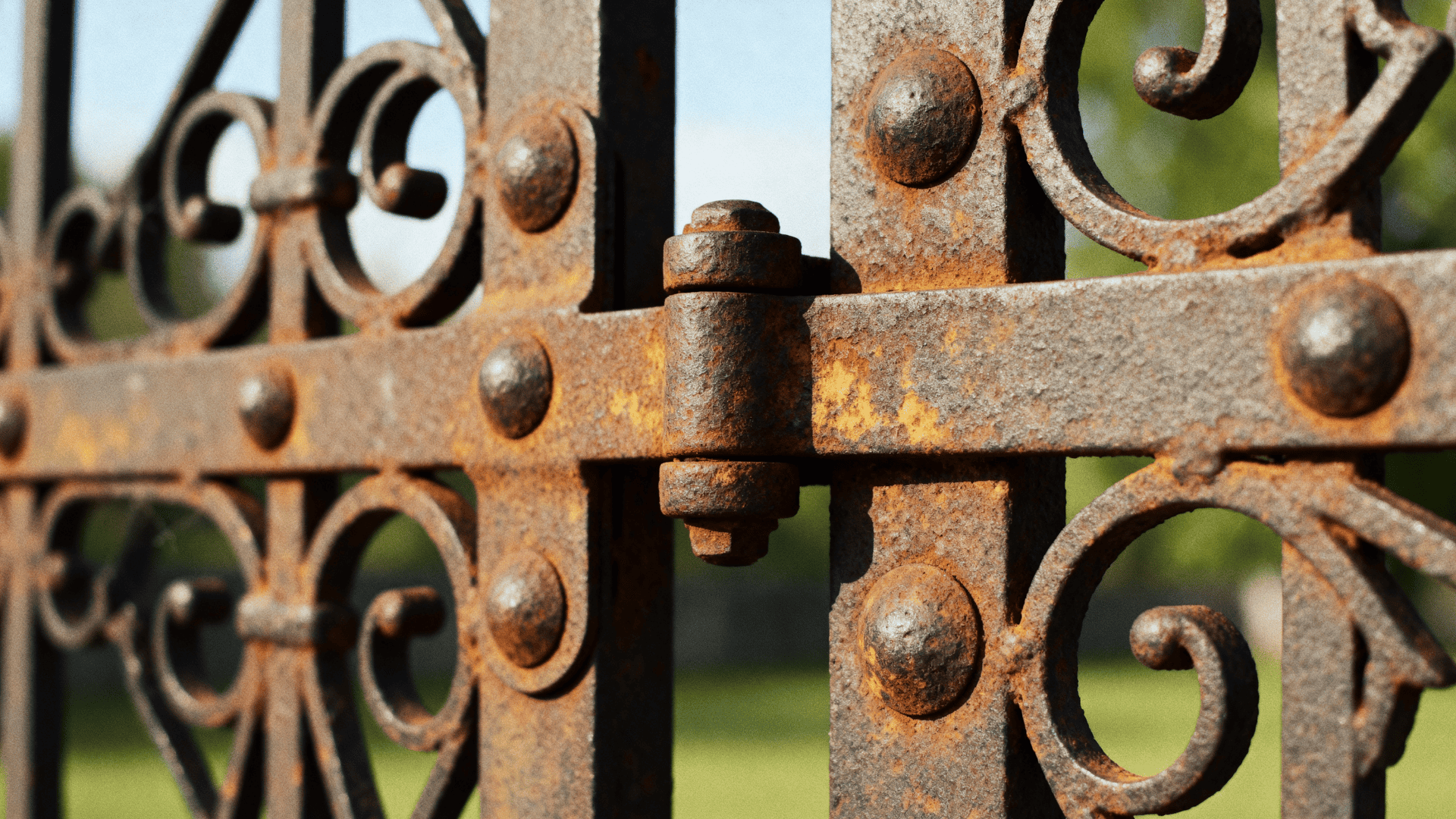 Close up of a rusty wrought iron gate hinge with decorative scrollwork, showing weathered metal texture and bolts in natural light.