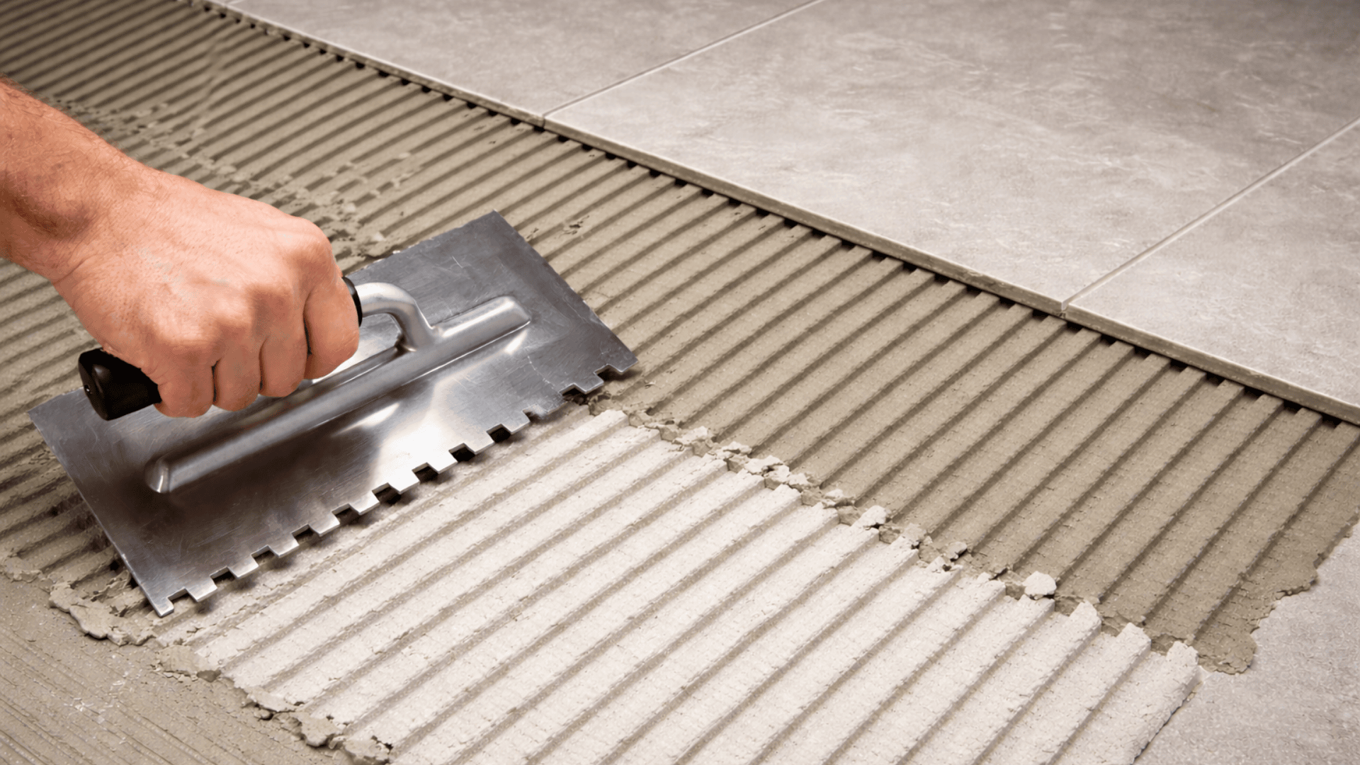 Close-up of a square-notch trowel spreading thinset mortar in straight ridges for installing large 12x24 floor tiles.