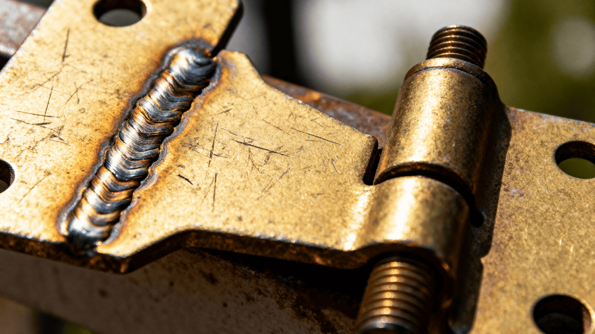 Close up of a welded metal hinge with visible weld bead, scratches, and threaded bolts, shown in bright outdoor light.