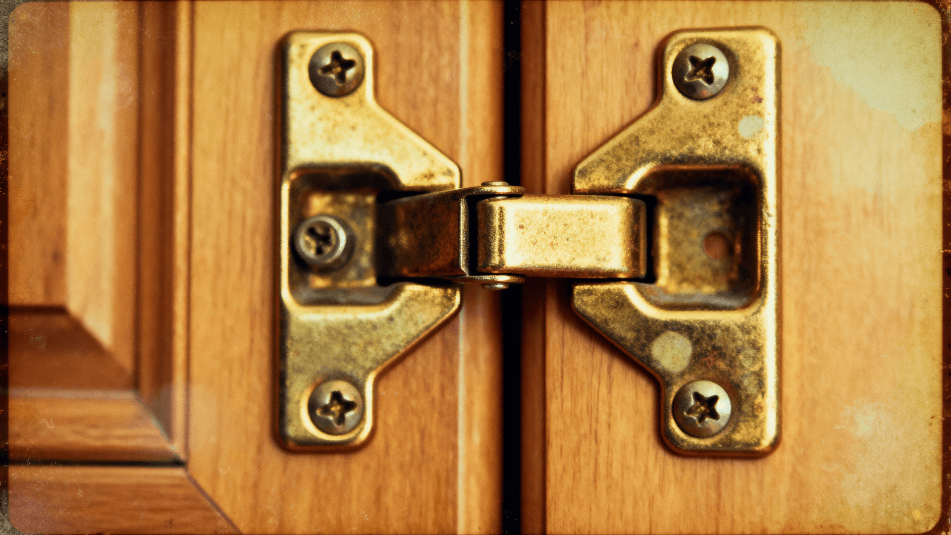 Close up of an overlay cabinet hinge mounted on wooden doors, showing brass finish, screws, and hinge arm connecting the panels.