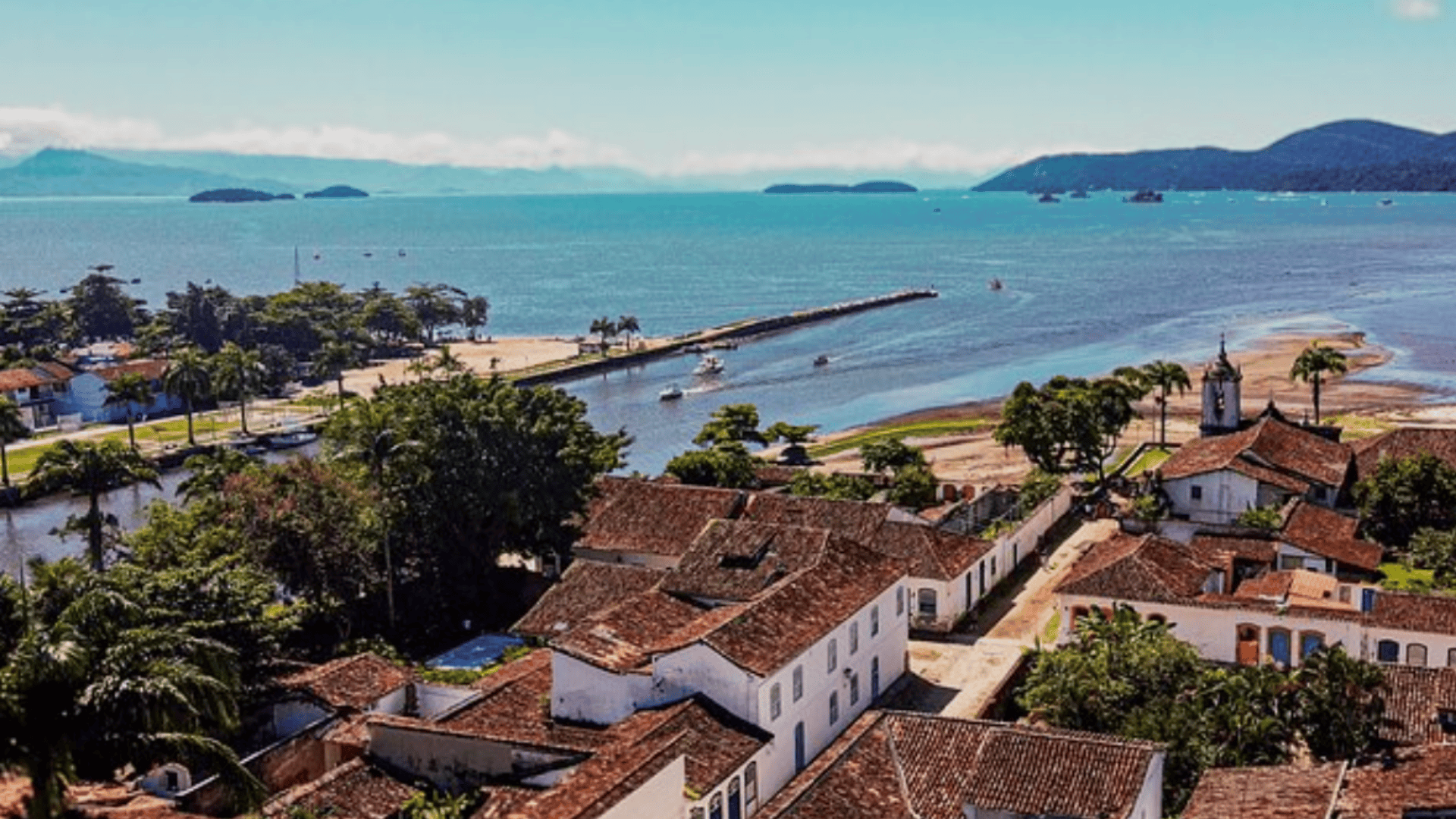 Coastal village with red tiled roofs overlooking a blue bay, sandy beach, and distant islands under a clear sky.