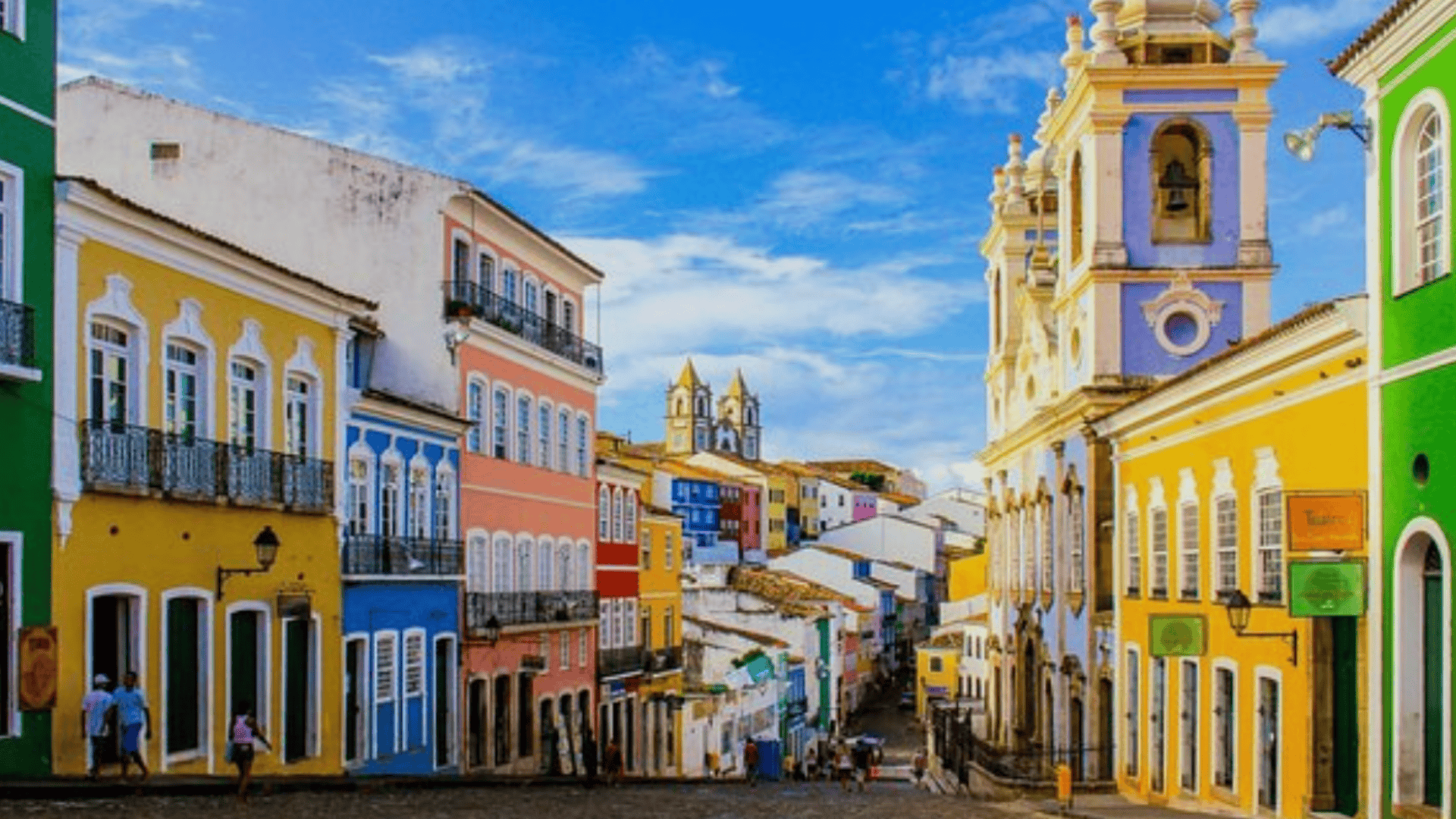 Colorful colonial street in Salvador, Brazil, with pastel buildings, cobblestones, and a baroque church tower under a bright blue sky.