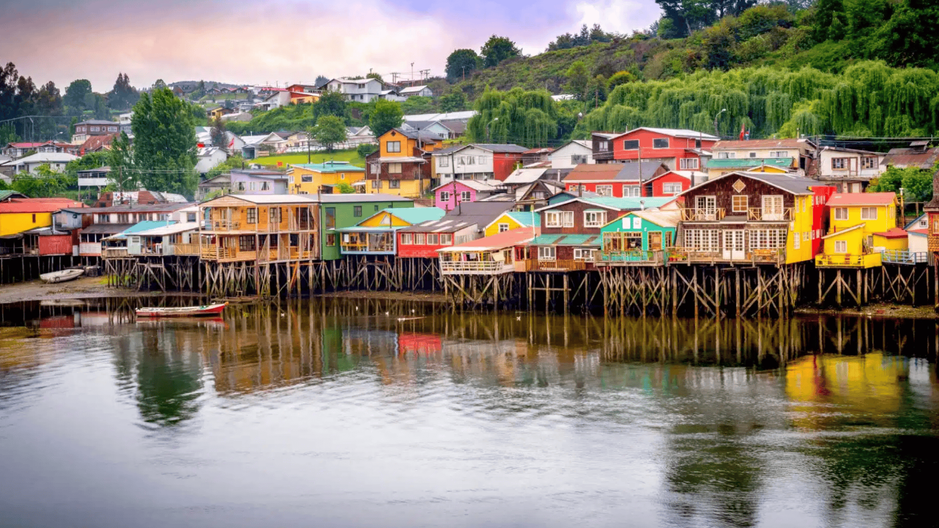 Colorful stilt houses line a calm waterfront, reflecting in the water, with green hills and scattered homes under a soft sky.