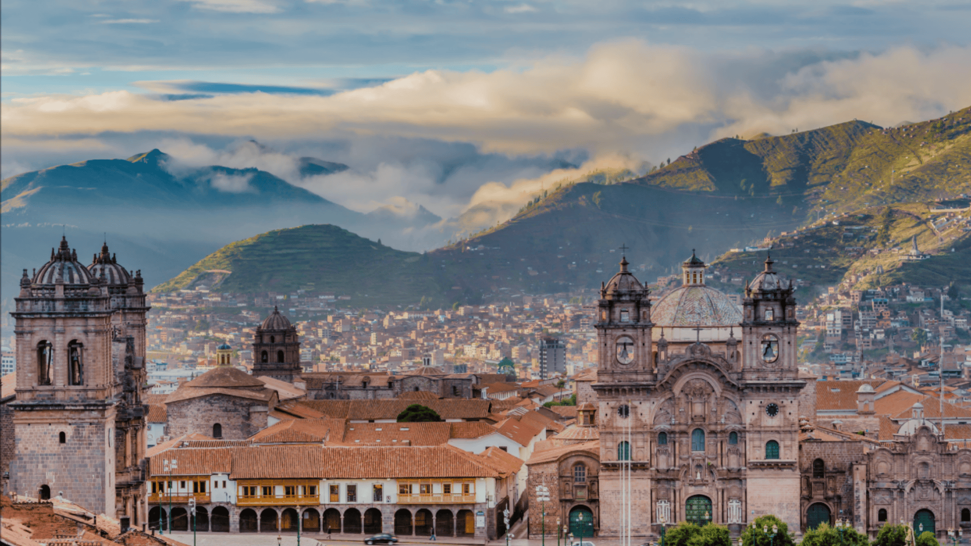 Cusco cityscape with historic cathedral and stone buildings, red rooftops below green Andean hills and low clouds at sunrise.