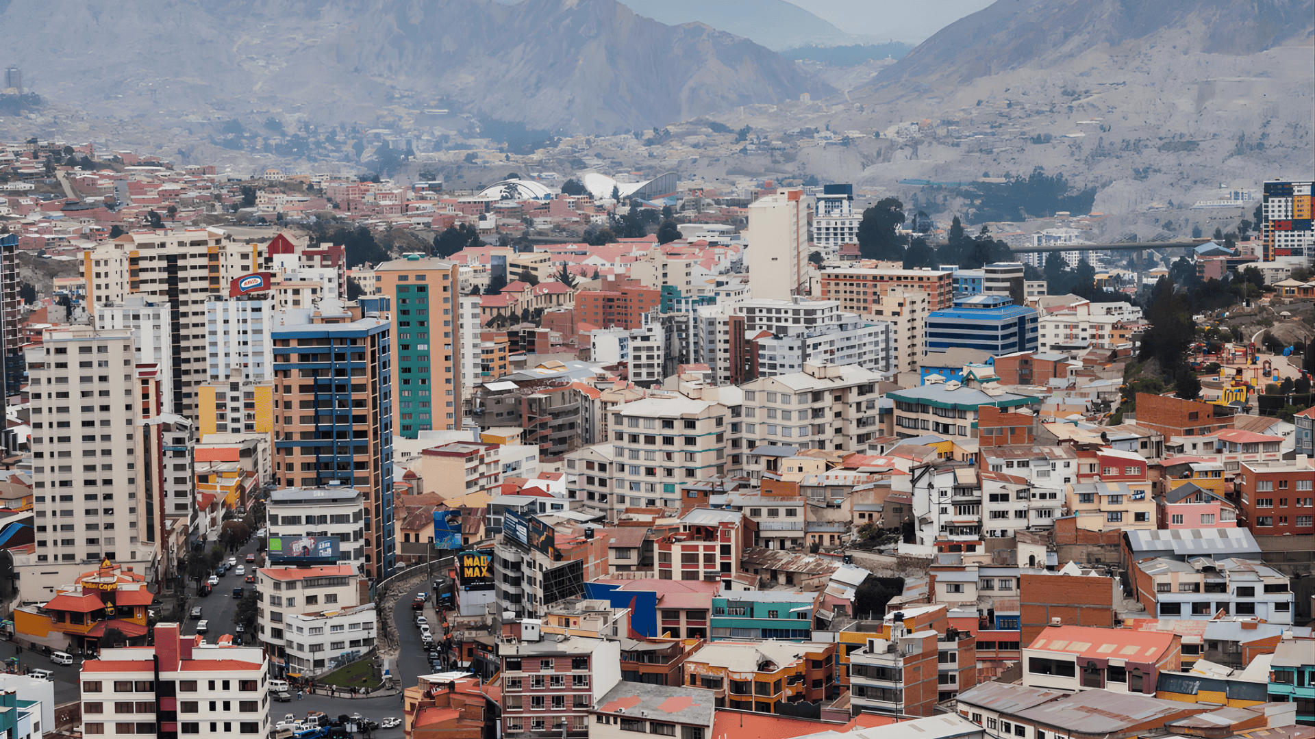 Dense cityscape with mixed high rises and low buildings spread across a valley, backed by rugged mountains under an overcast sky.
