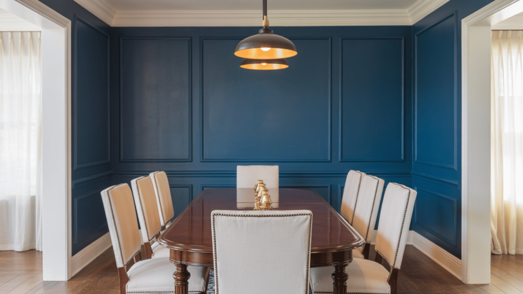 Dining room with deep navy accent wall, paneled trim, wood table, cream chairs, and warm pendant lighting.