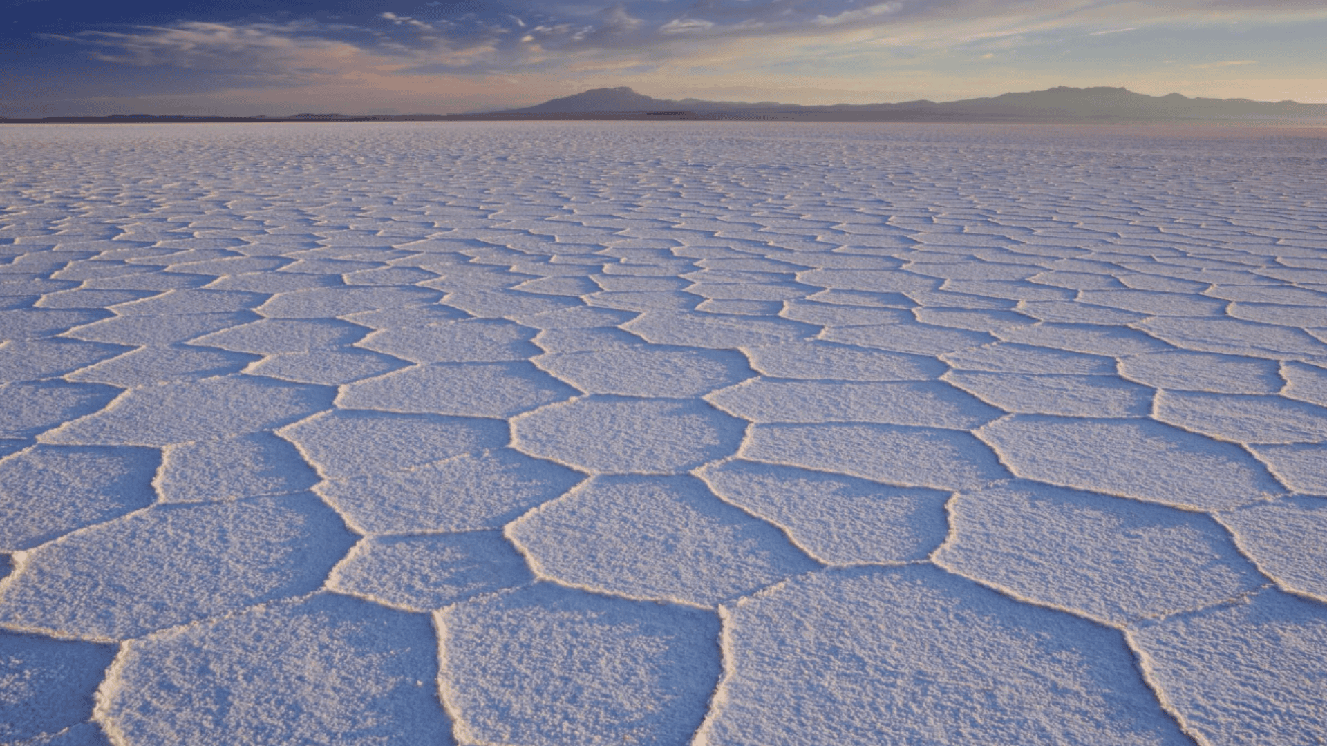 Endless salt flat with hexagonal crust patterns, fading into distant mountains under a pastel sky at sunrise or sunset.