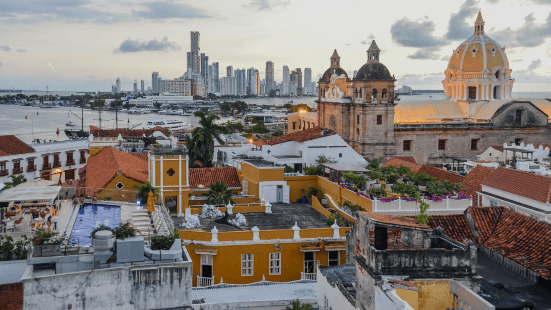 Historic coastal city with colorful rooftops and a large cathedral dome, overlooking the sea and a modern skyline in the distance.
