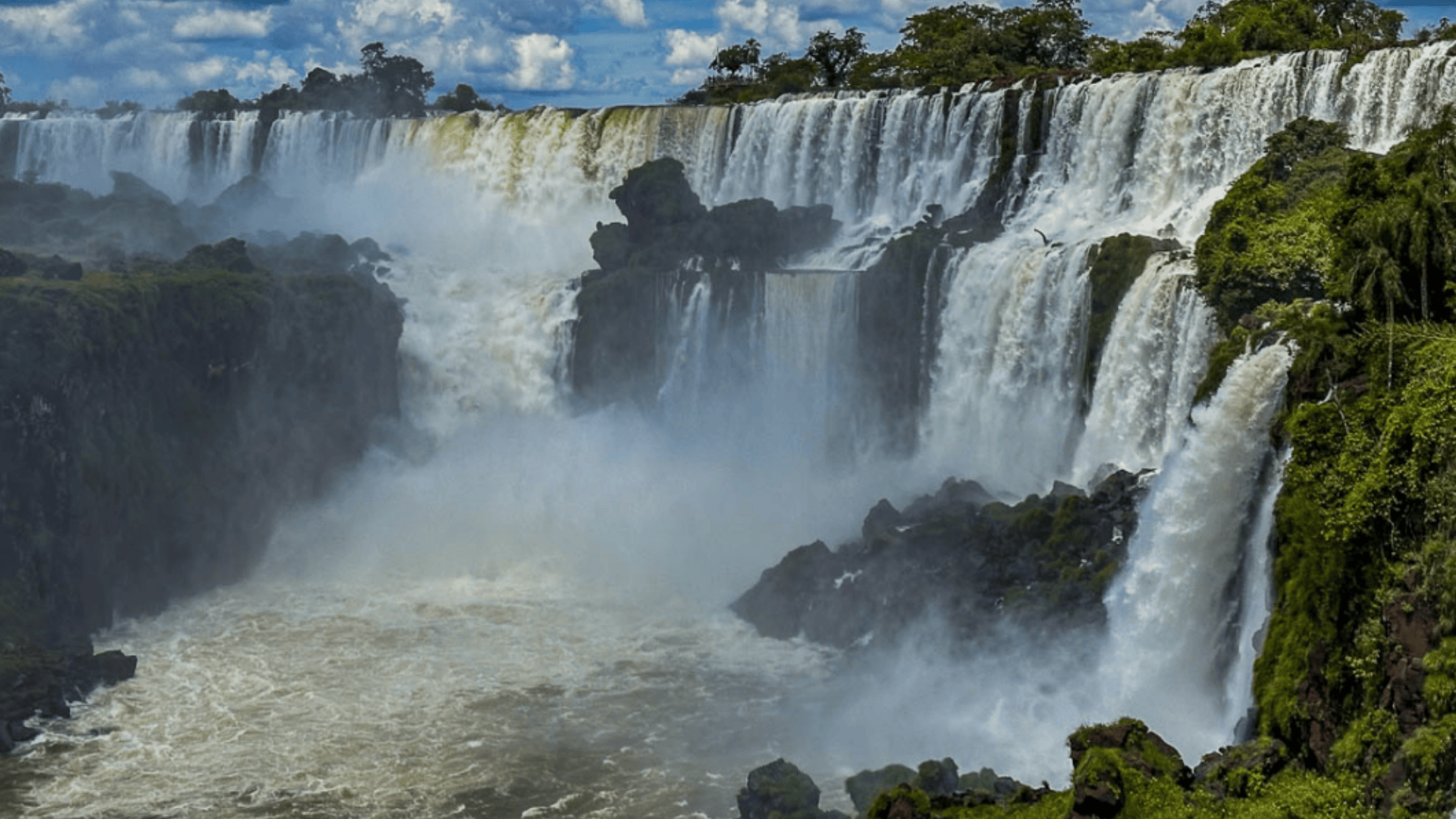 Iguazu Falls cascading over wide cliffs, powerful waterfalls surrounded by lush green rainforest under dramatic clouds.