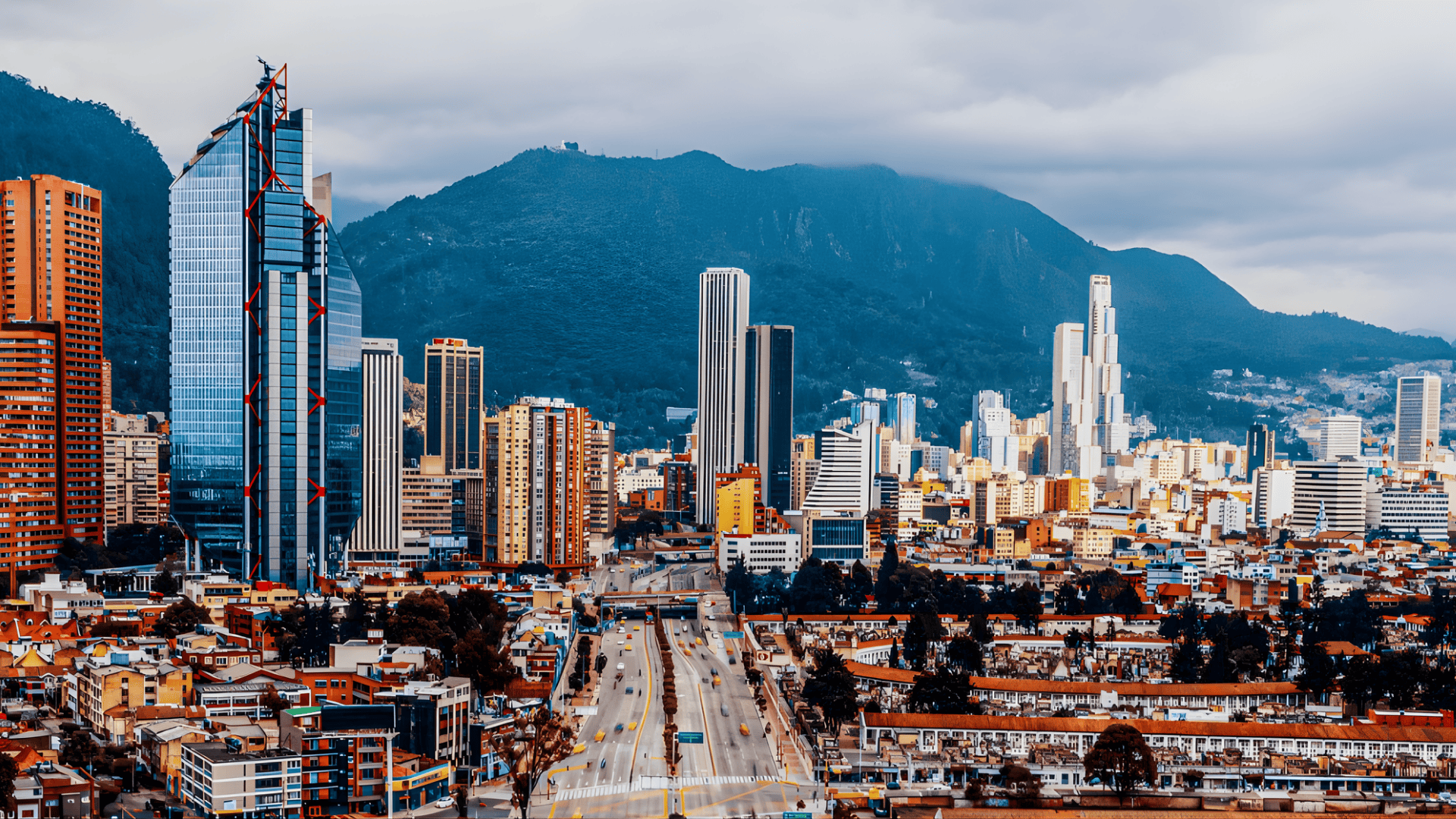 Modern city skyline with tall glass skyscrapers and busy roads, set against dark green mountains under a cloudy sky