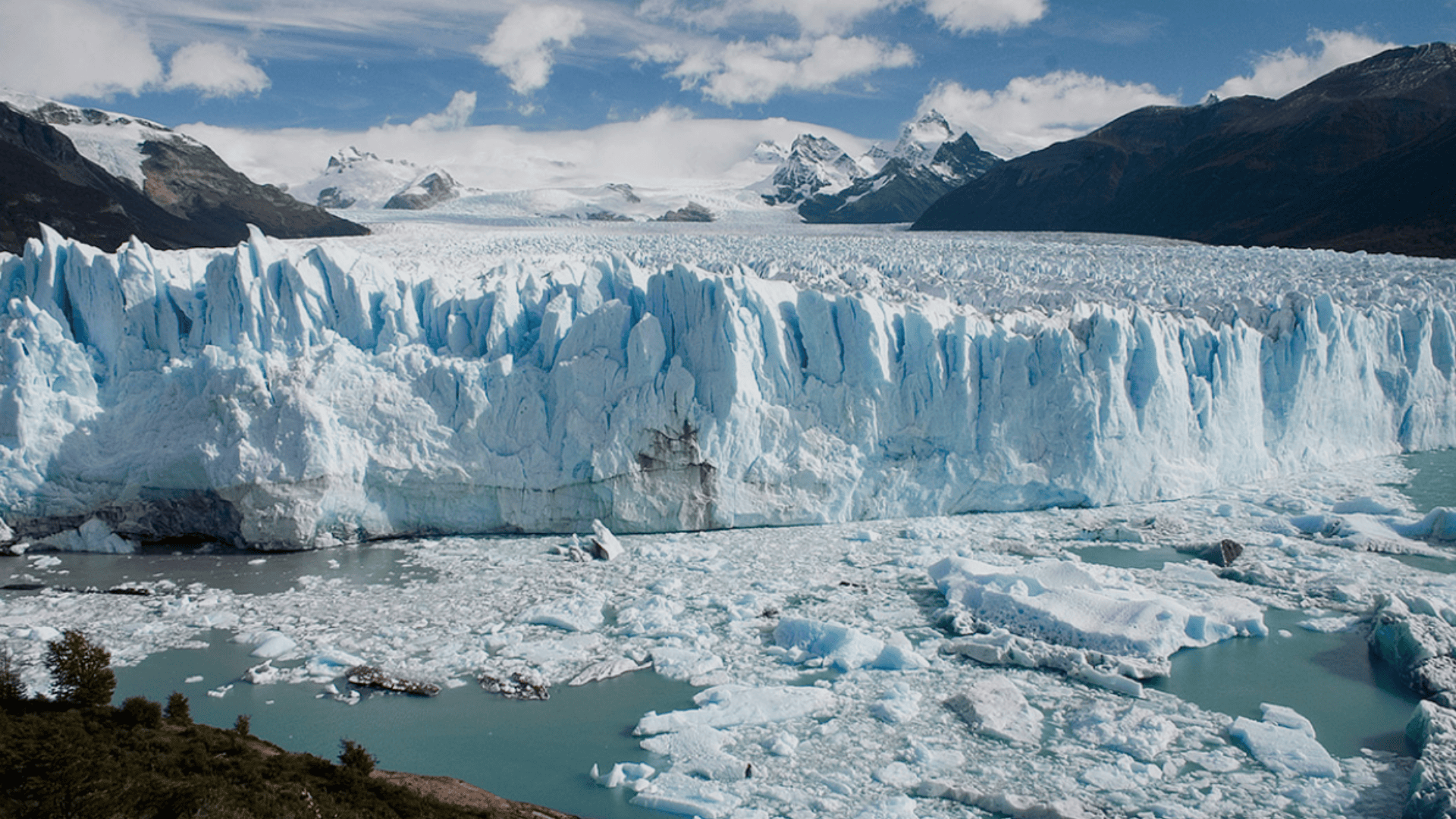 Perito Moreno Glacier with towering blue ice wall, floating ice chunks in turquoise water and rugged mountains under a partly cloudy sky.