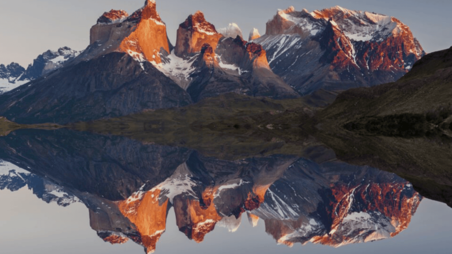 Snow capped mountain peaks glow at sunrise, reflected perfectly in a calm lake, creating a symmetrical landscape under a clear sky.