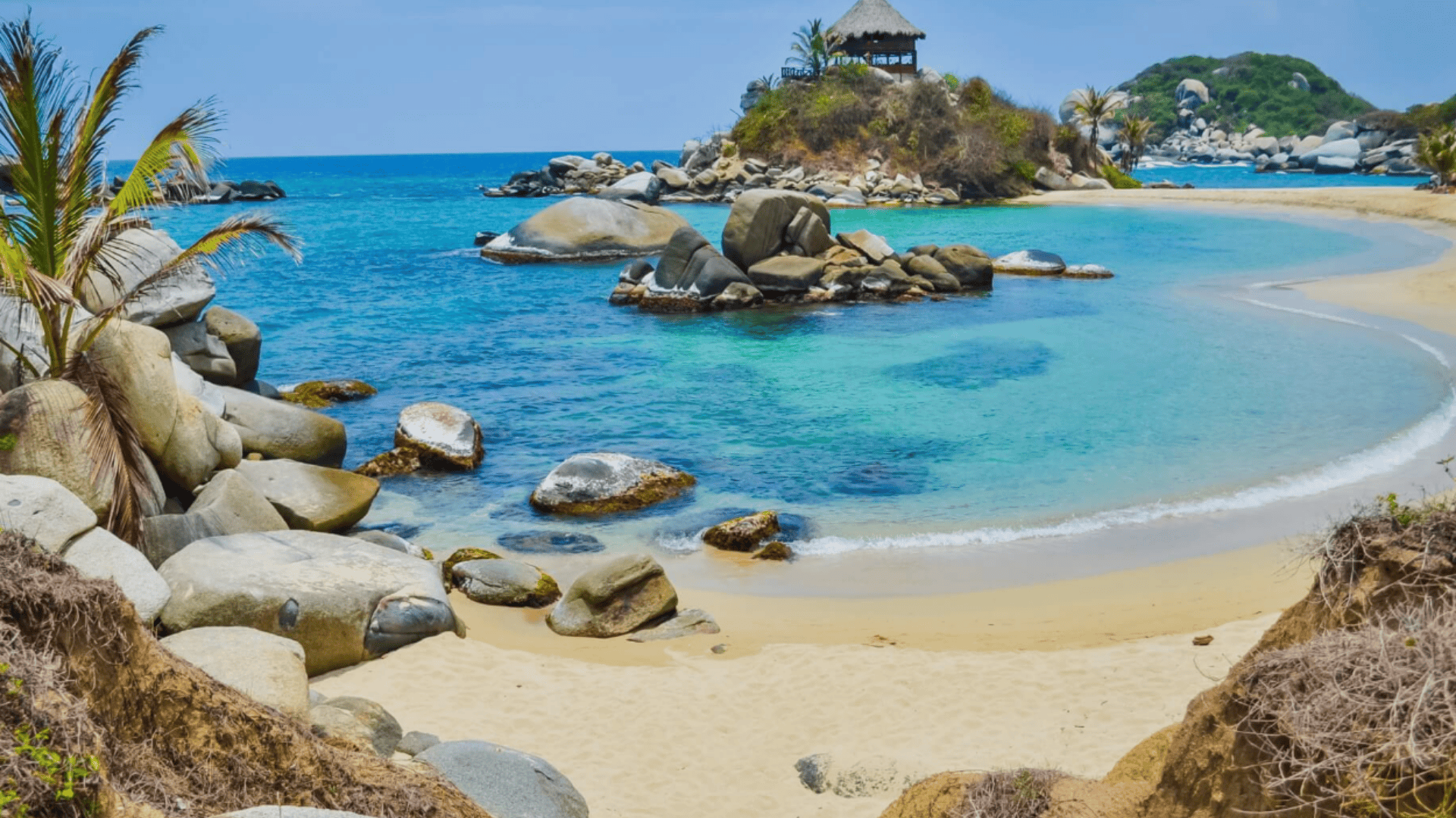 Tropical beach with golden sand, turquoise water, and large boulders, with palm trees and a small rocky island in the distance.