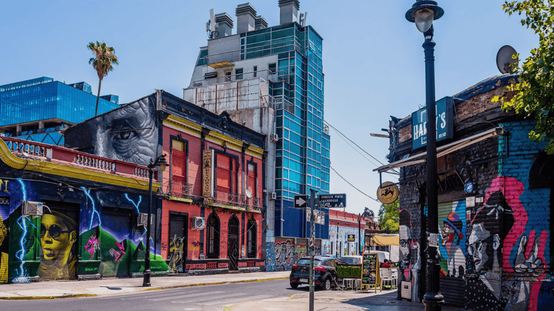 Urban street with colorful murals on buildings, a tall glass tower in the background, and bright daylight over a quiet road.