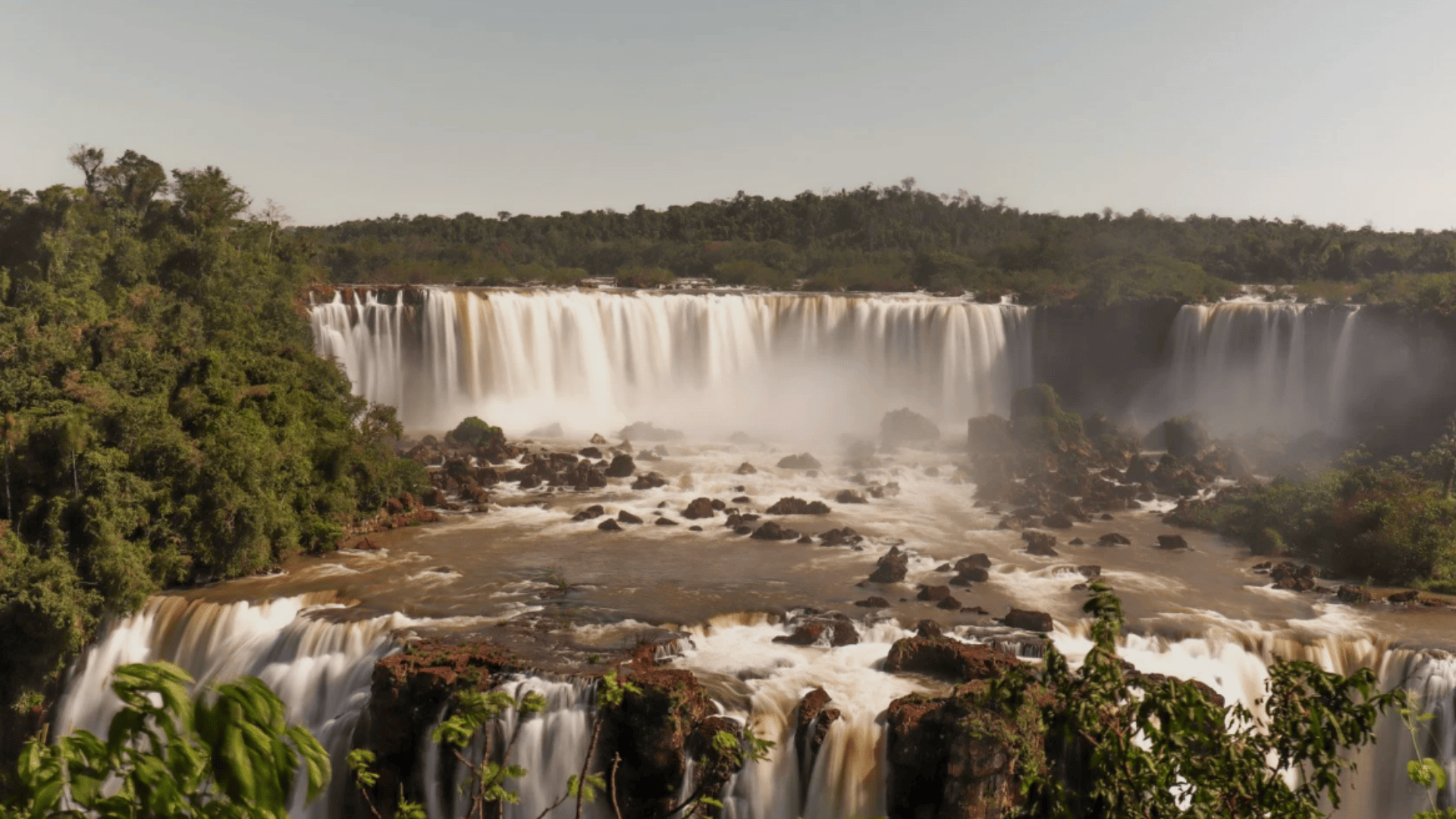 Wide view of Iguazu Falls pouring over cliffs into misty river, surrounded by dense green rainforest under a clear sky.