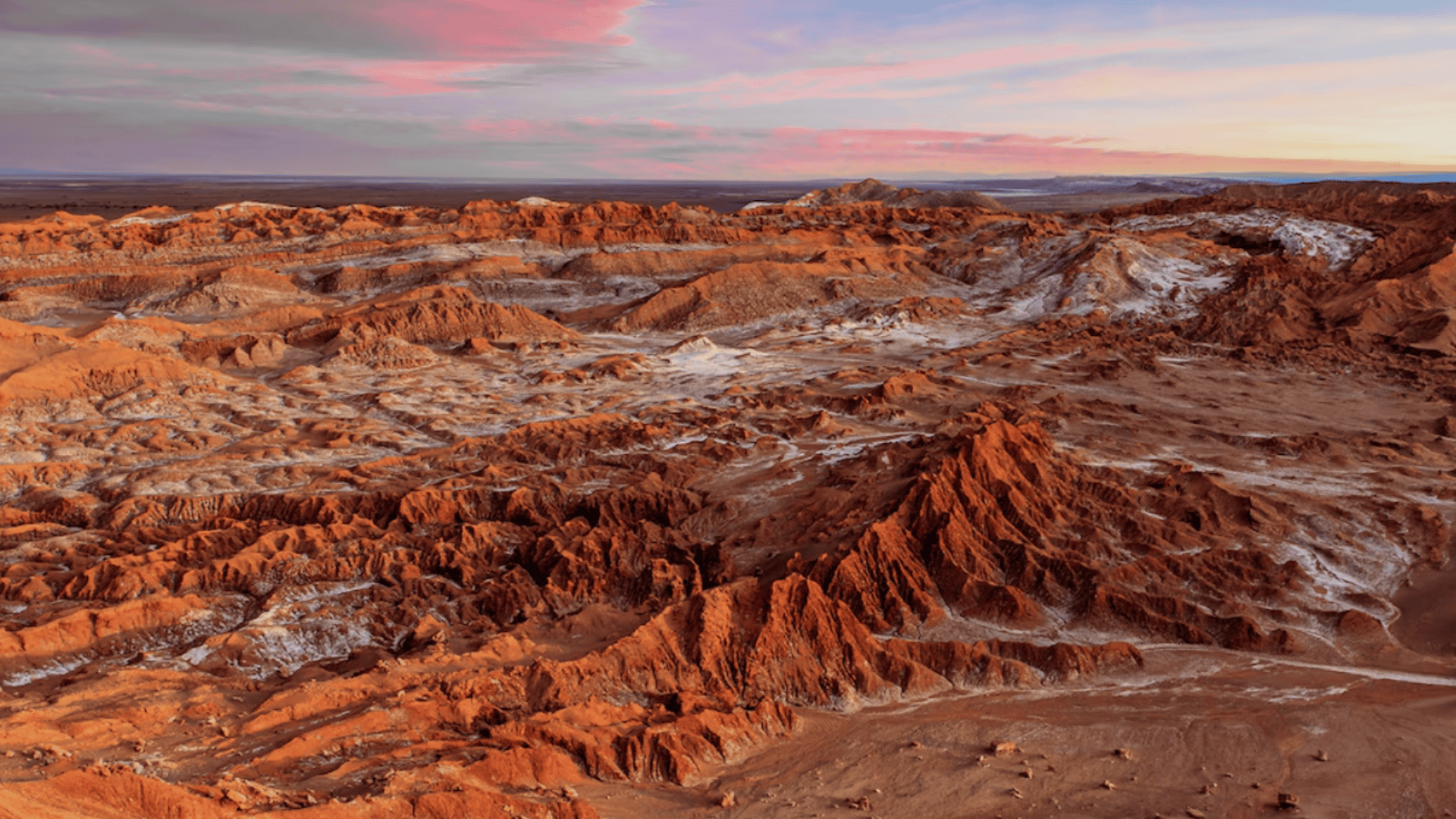 Wide view of a rugged red desert with rocky ridges and salt flats, glowing under a pink and orange sunset sky.