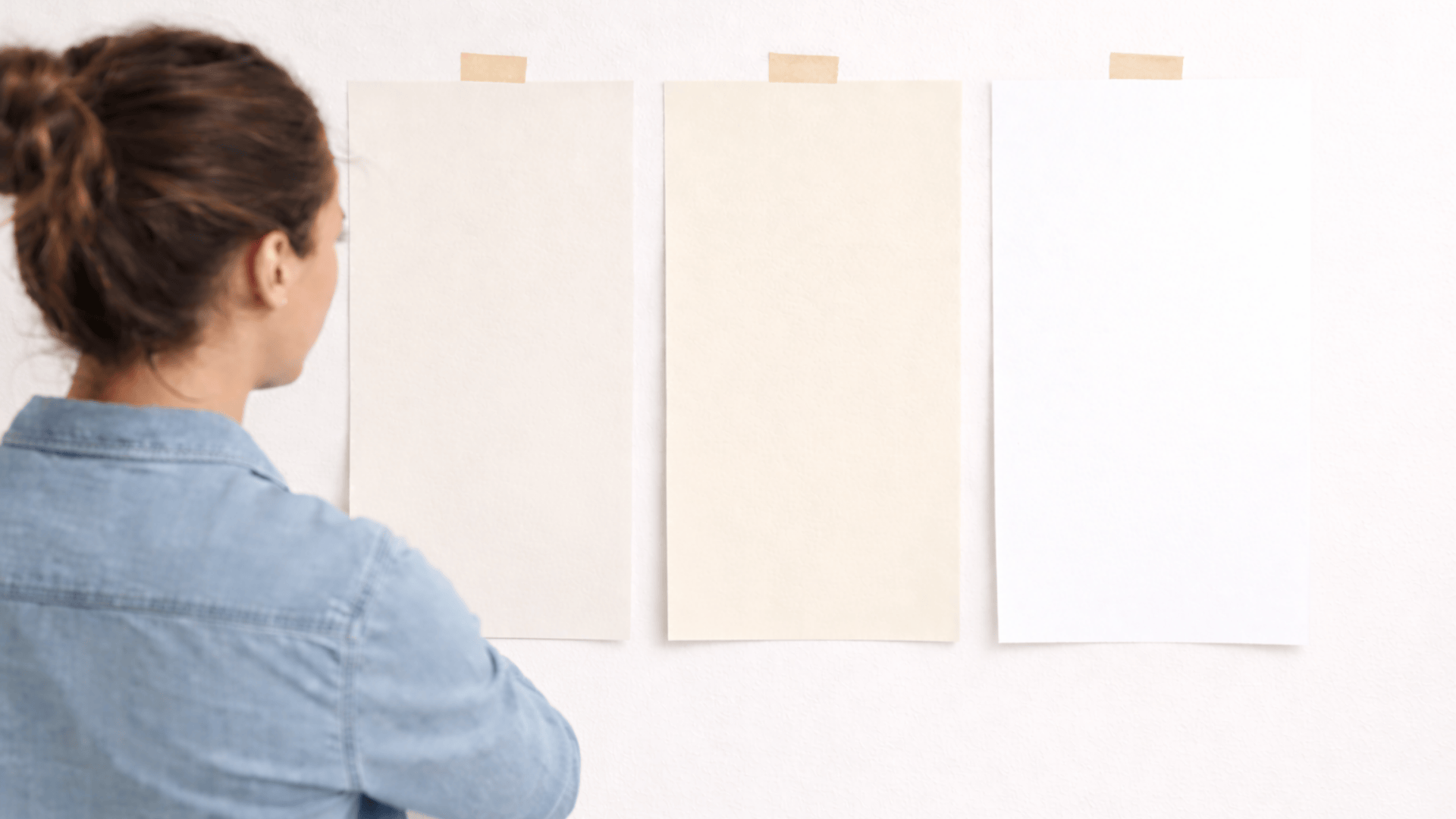 Woman compares three white paint swatches taped to a wall, showing subtle warm-to-cool shades in bright natural light.