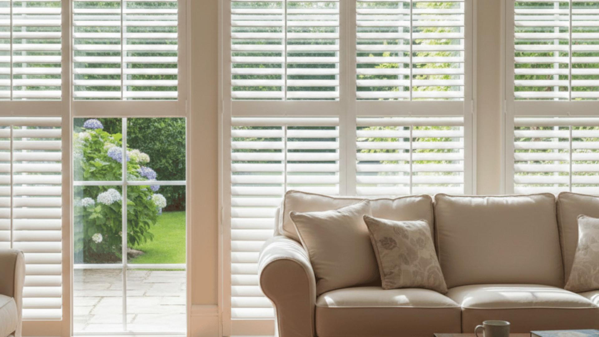 a bright living room with white plantation shutters, a beige sofa, and a wooden coffee table, overlooking a garden outside.