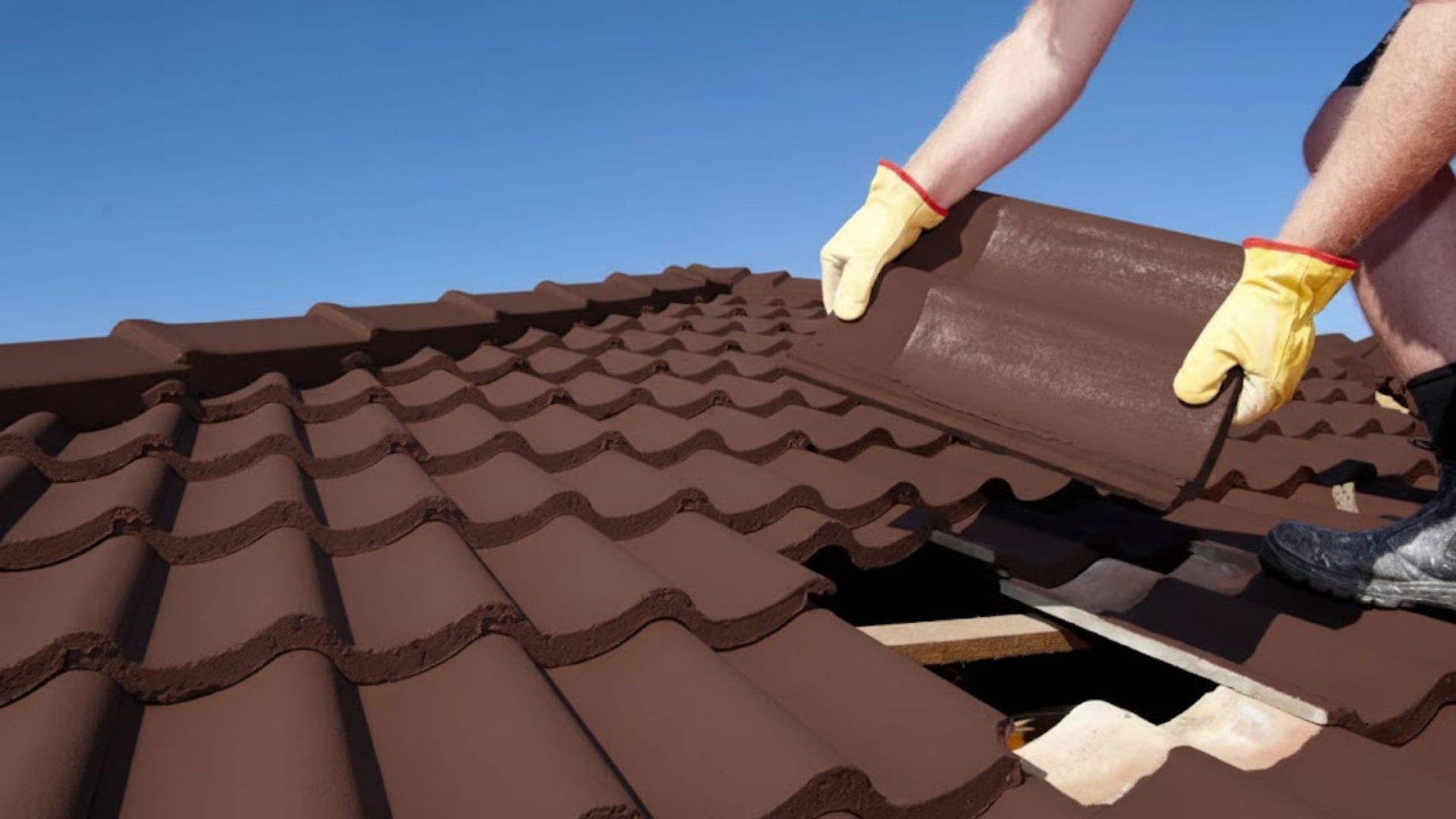 a roofer in yellow gloves installs dark brown terracotta tiles on a sloped roof under a clear, bright blue sky