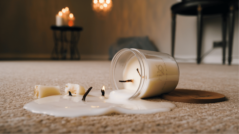 a spilled candle on a beige carpet, with wax flowing out of a glass jar and a couple of broken candle pieces nearby. The candle flame is still lit