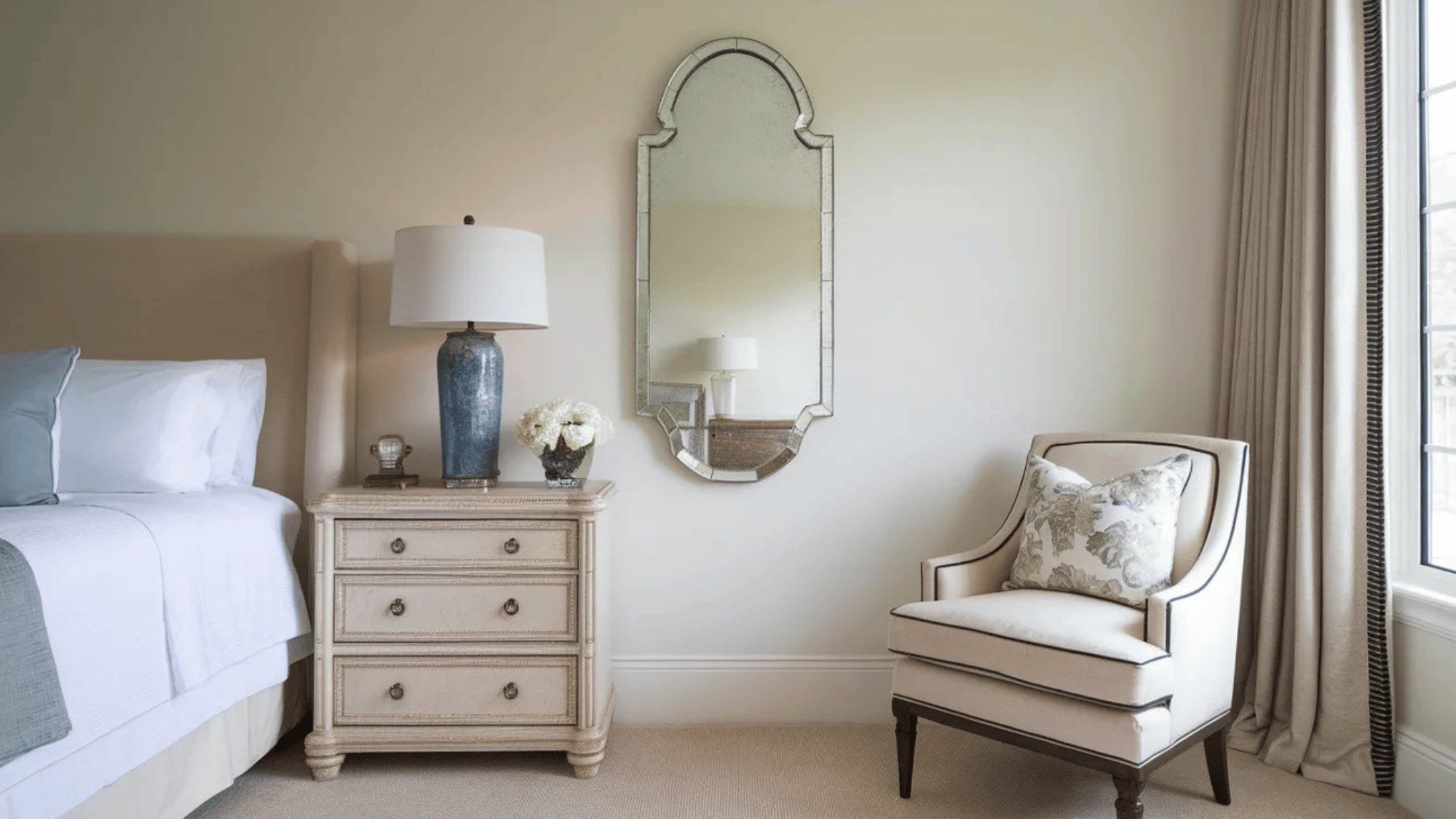a stylish bedroom corner featuring a decorative wall mirror above a beige nightstand with a blue vase and white flowers, next to a plush armchair