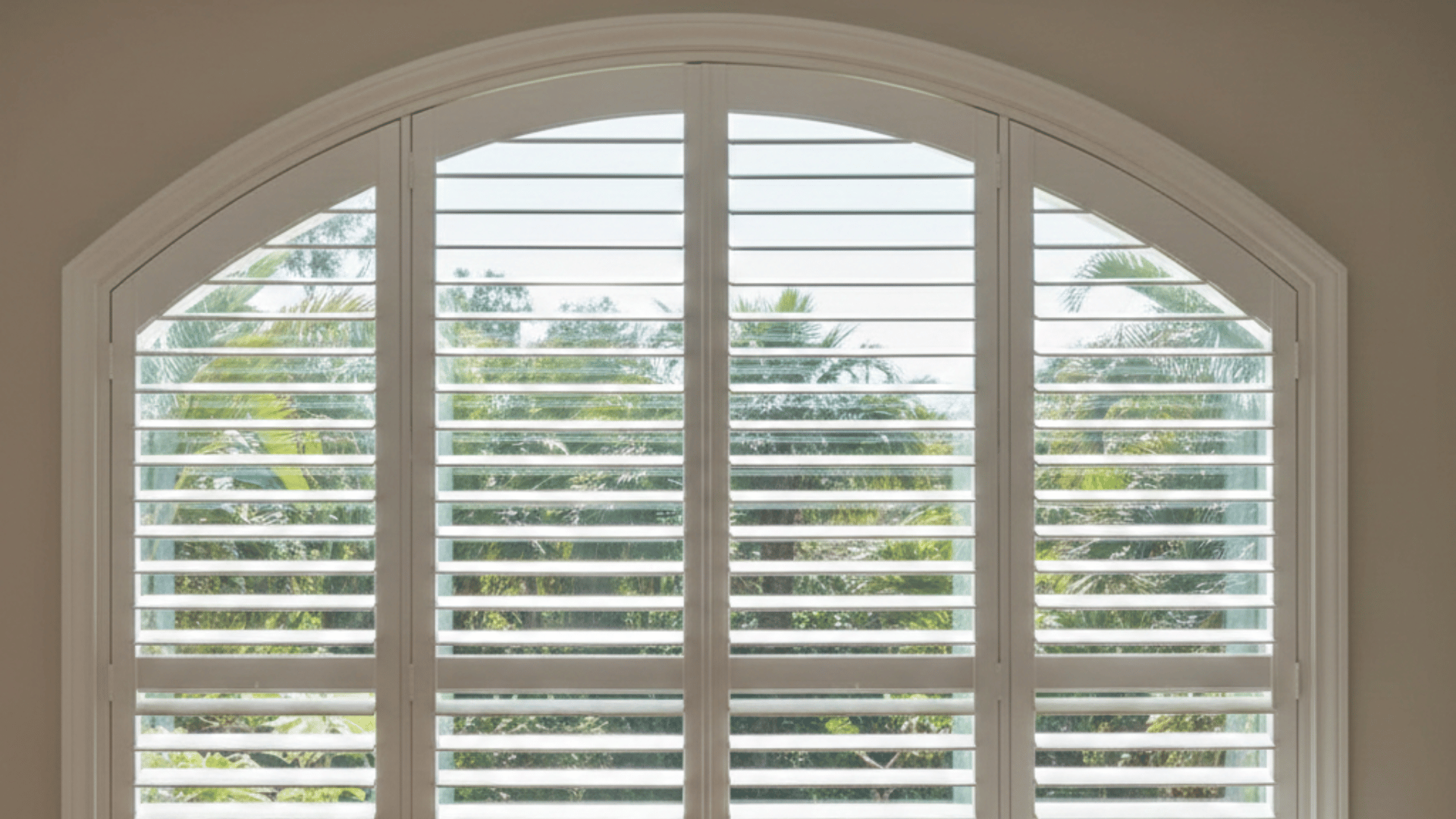 a well-lit room featuring white plantation shutters with an arched window and a cozy armchair beside it, casting soft shadows on the floor.