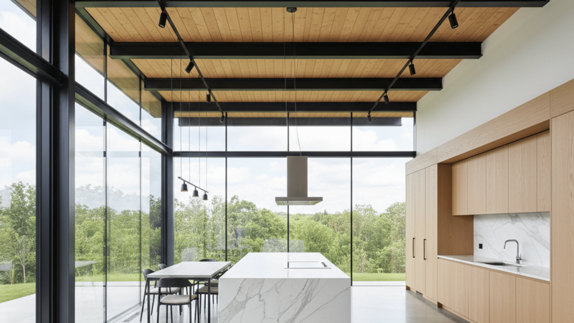 modern kitchen with a light wood ceiling, black beams, marble island, and floor-to-ceiling windows showing green trees.