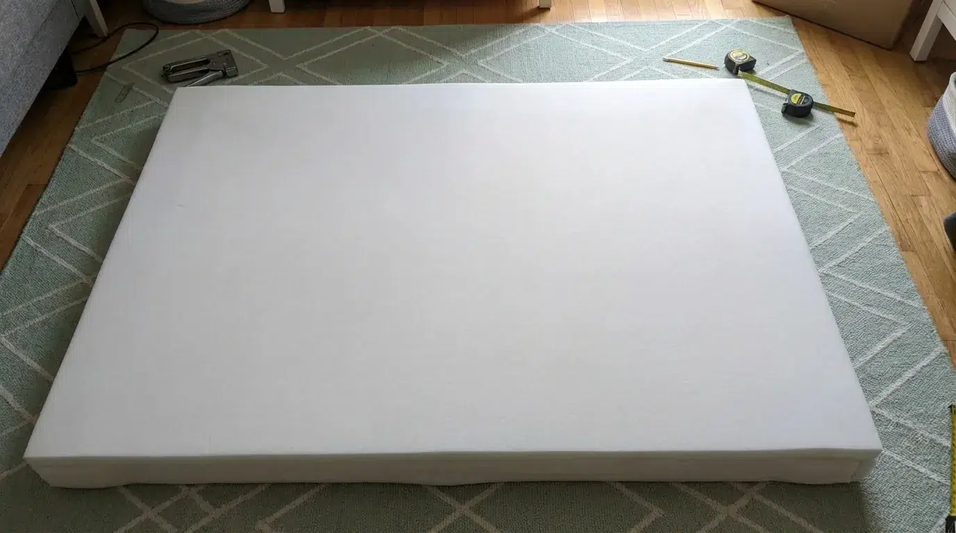 Overhead view of thick white foam being placed onto a plywood headboard base on a green patterned rug during a DIY upholstered headboard build