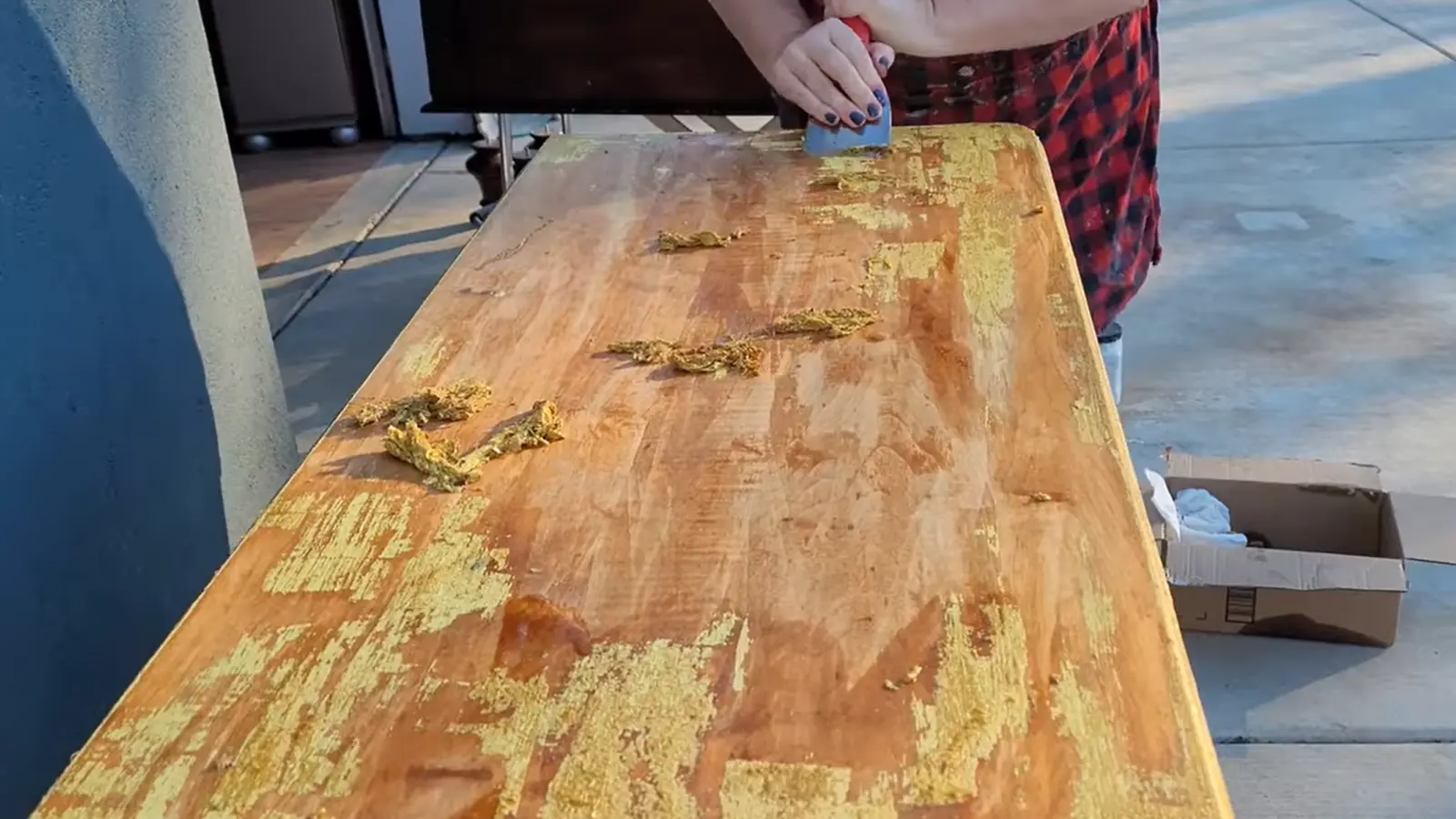 Person scraping thick yellow finish from a wooden dresser top using a putty knife after applying chemical stripper.