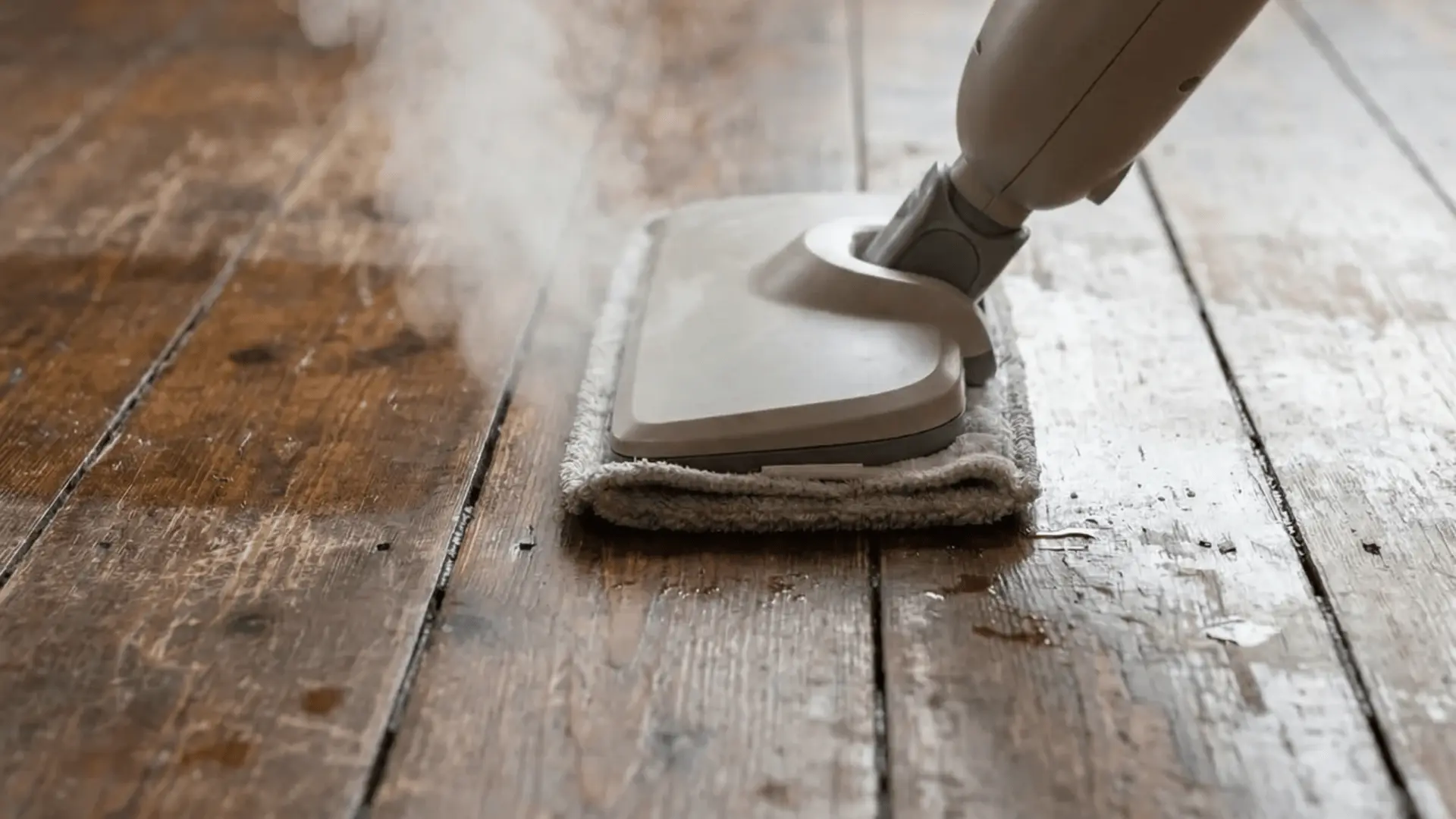 Steam mop releasing steam onto hardwood floor boards with moisture visible between plank gaps