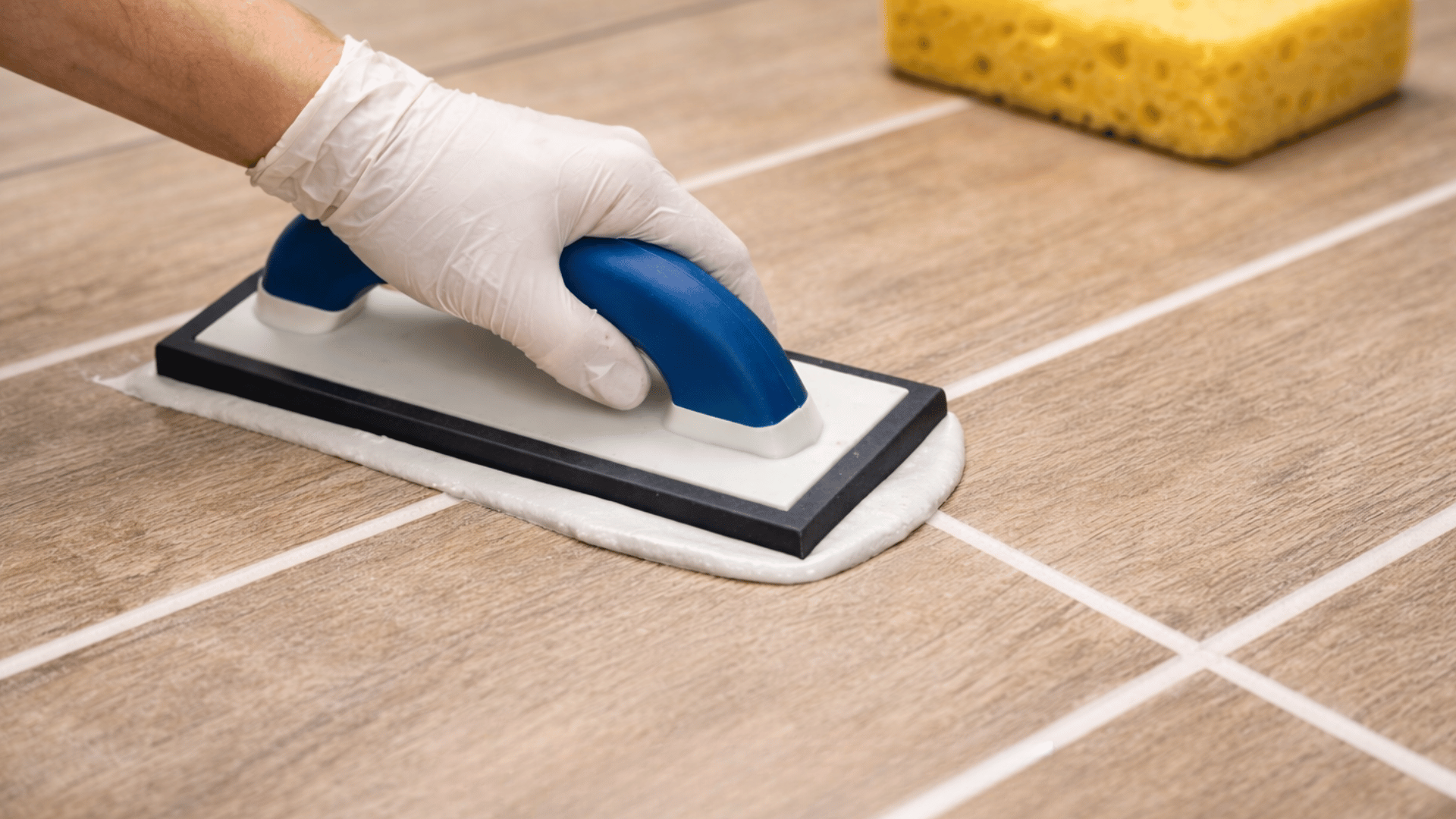 gloved hand uses a blue float to apply grey urethane grout to wood-look tiles. a yellow sponge sits in the back