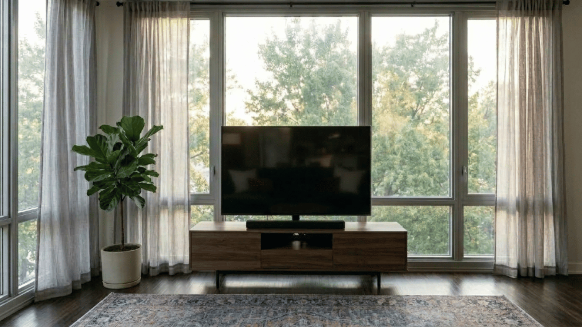 living room with a tv placed in front of tall windows, light curtains, and warm natural light