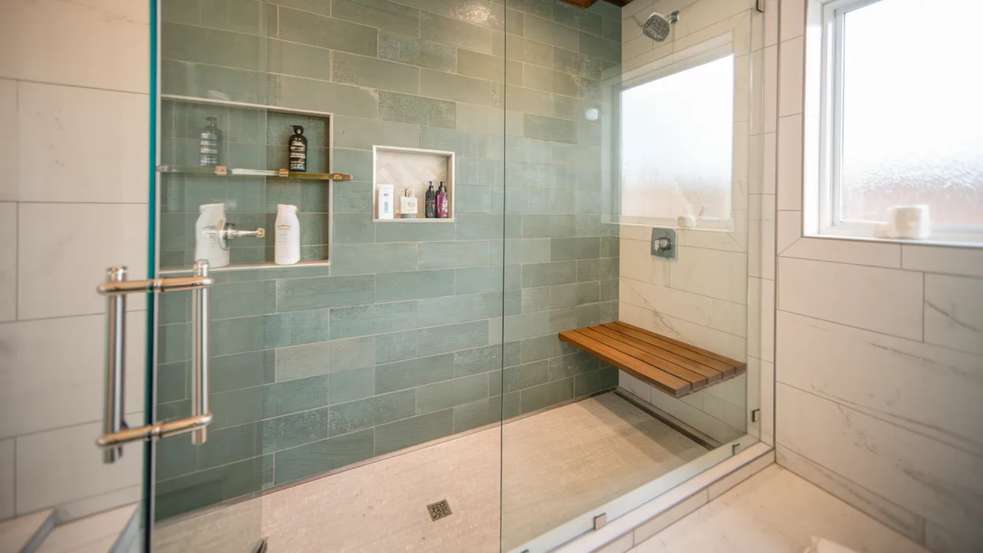 modern shower with glass enclosure green tile wall built in shelves wooden bench rainfall showerhead and window letting in natural light