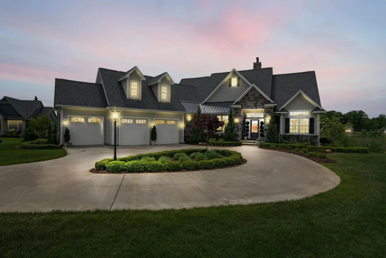 Large modern farmhouse with stone and siding exterior at dusk, circular driveway, and manicured shrubs