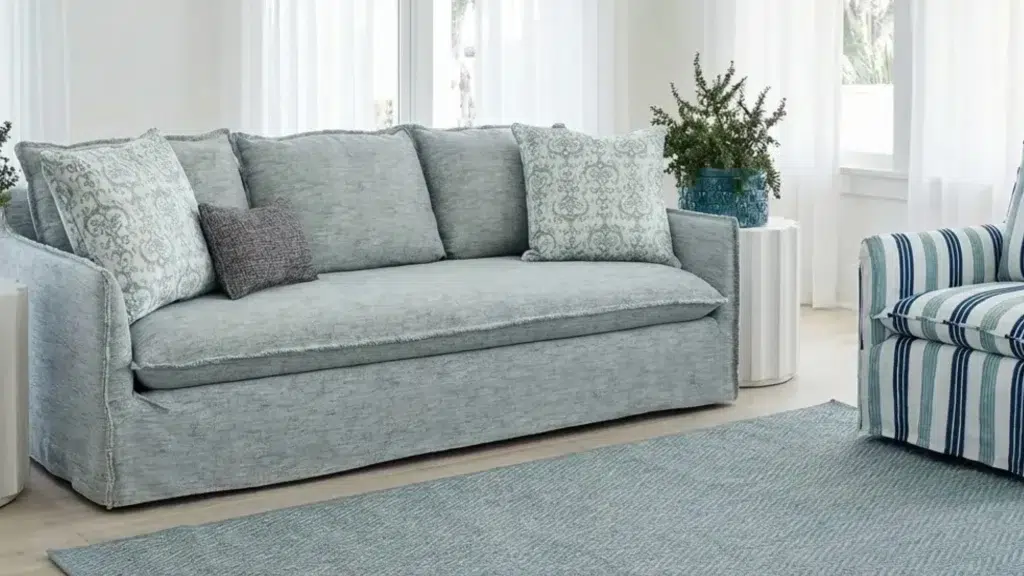 Coastal living room with a grey-blue linen slipcover sofa, sheer curtains, white walls, and soft blue accents.
