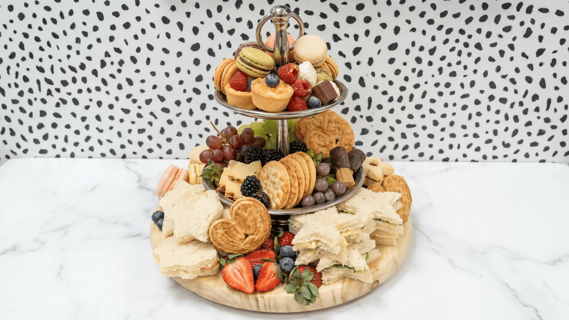 A tea-time board with biscuits, mini sandwiches, fruit, jam, butter, and sweet treats arranged in small portions for a light afternoon gathering.