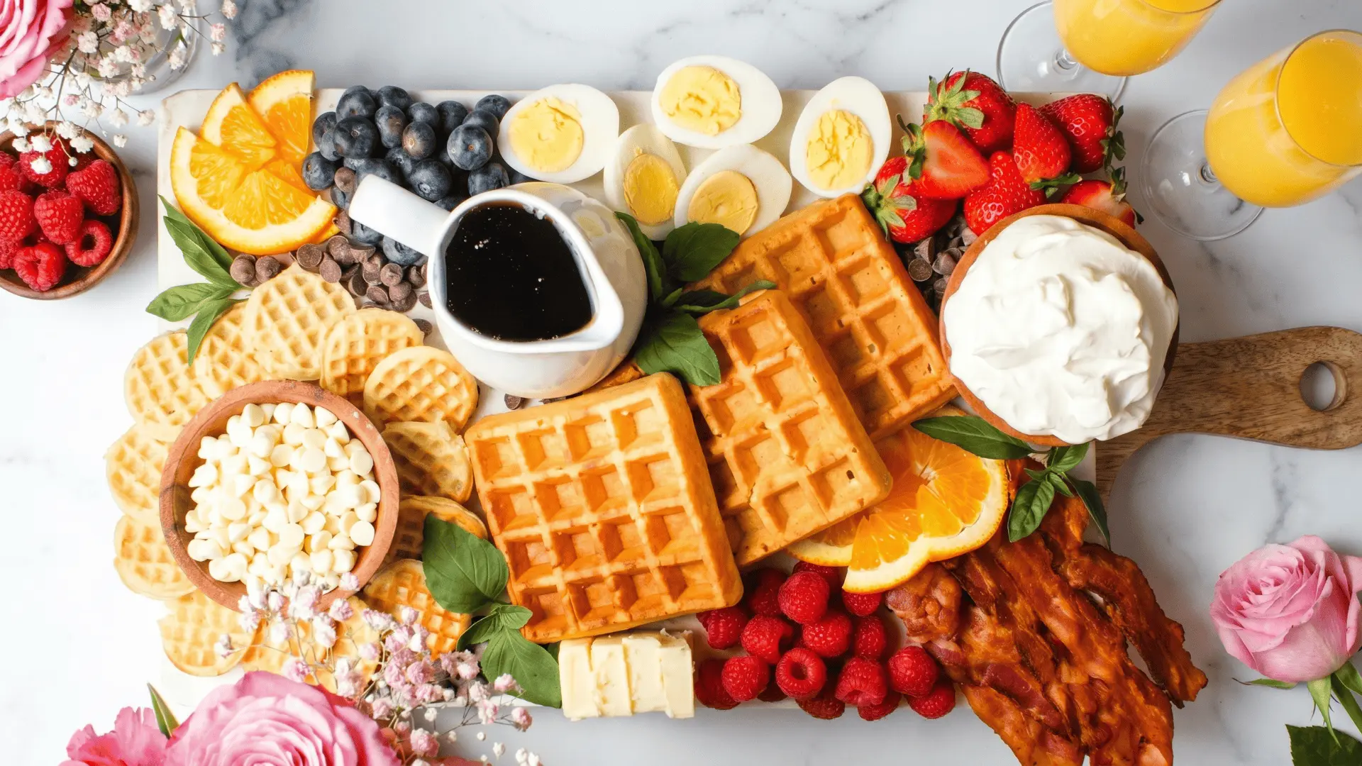 A waffle board with warm waffles, fresh berries, banana slices, syrup, honey, chocolate spread, whipped cream, nuts, and granola arranged for easy self-serve brunch.