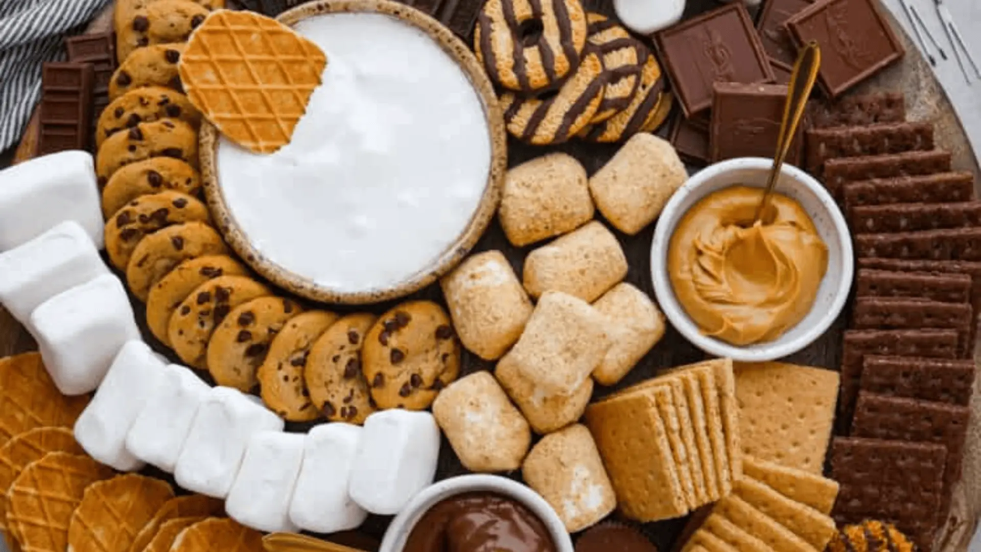 A wooden dessert board filled with marshmallows, cookies, graham crackers, chocolate bars, strawberries, and three dips ,chocolate, peanut butter, and marshmallow cream.
