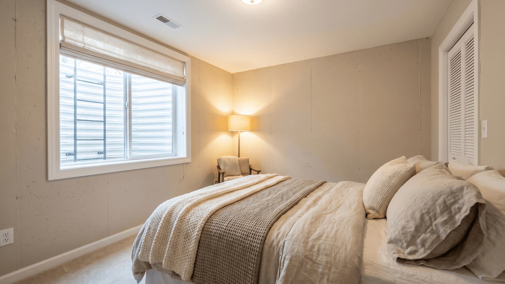 Basement guest bedroom with egress window, neutral bedding, warm lighting, and closet