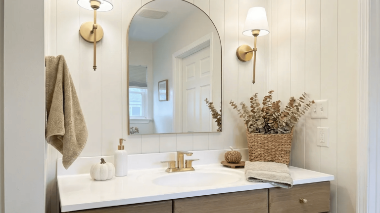 Bright neutral bathroom vanity with arched mirror, brass sconces, white countertop, woven decor, and folded towel