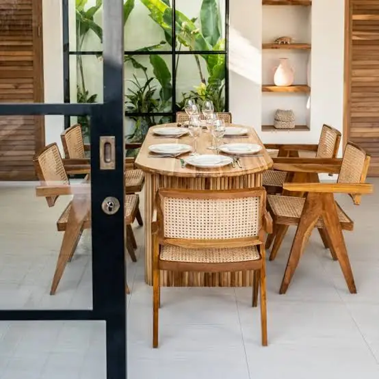 Wooden dining table with wicker chairs set for meal in airy room with large windows
