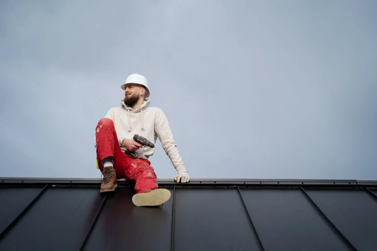 Construction worker in white hard hat and red pants sitting on metal roof holding drill