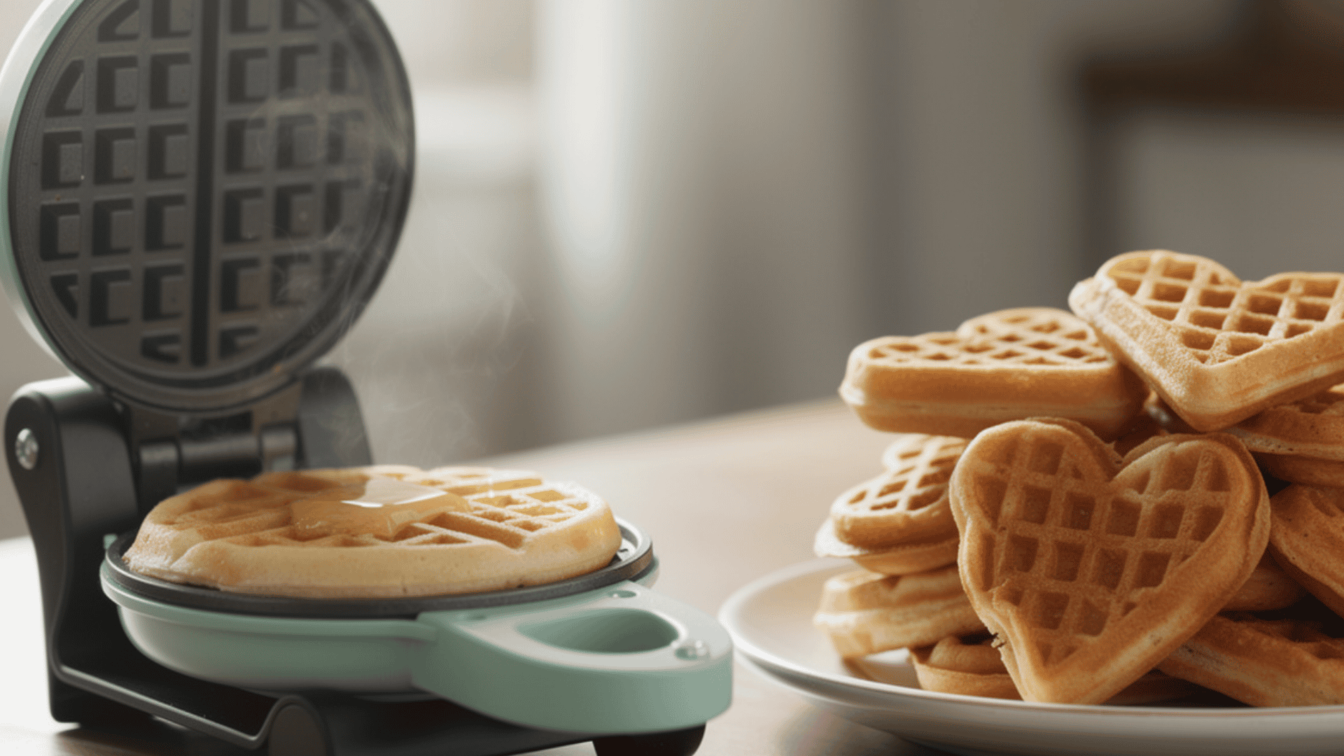 Mini waffle maker with a fresh waffle and a stack of heart-shaped waffles on a plate
