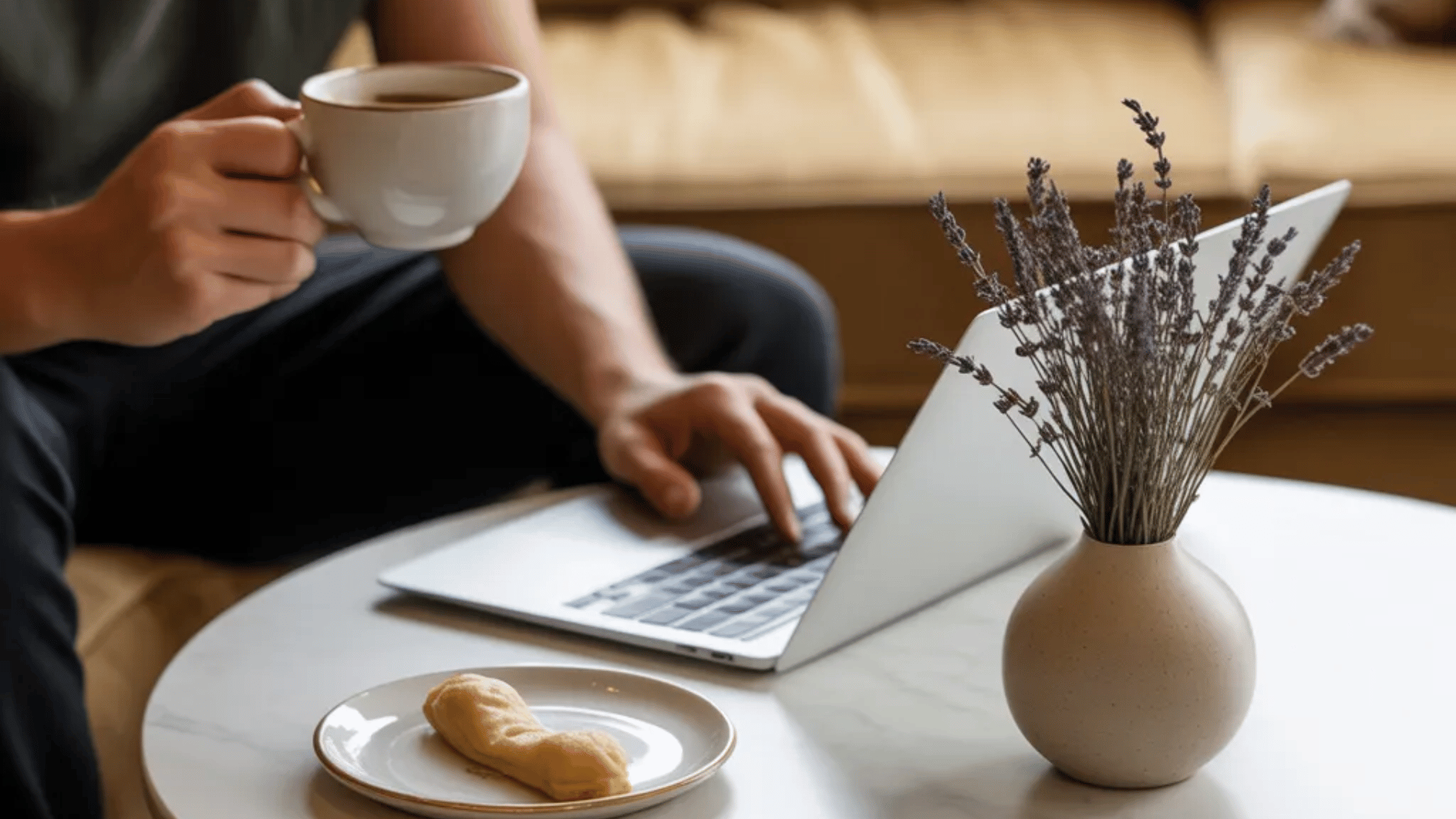 Person holding a coffee cup while using a laptop at a round table with a pastry and a vase of dried lavender.